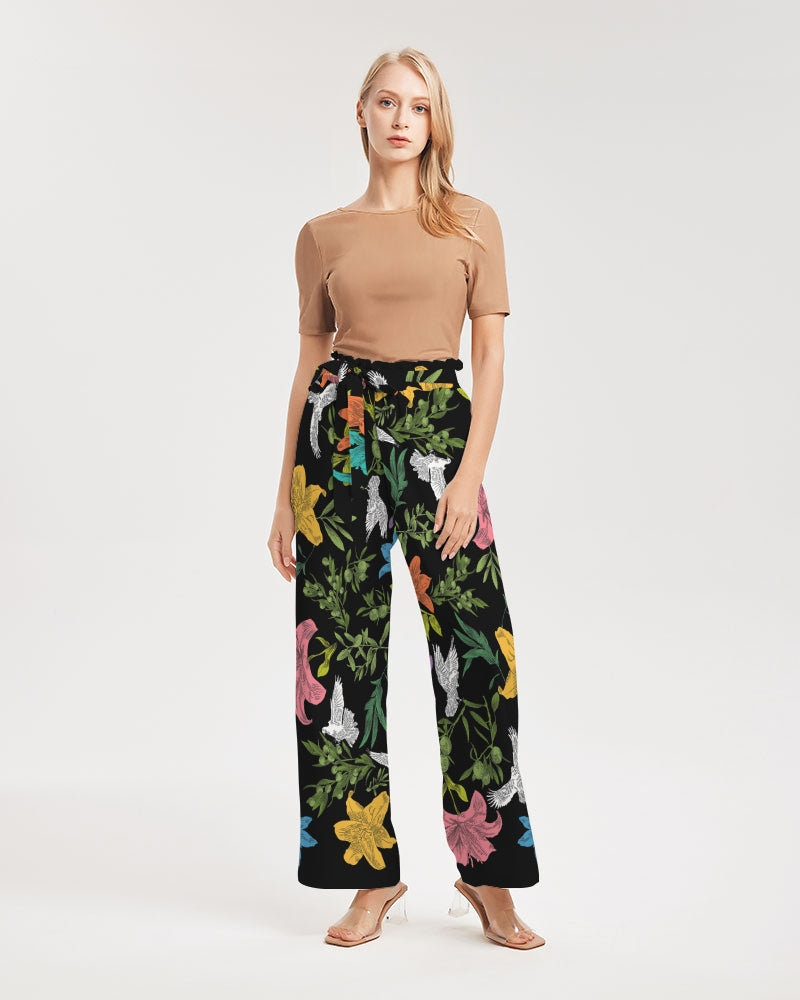 Front view of female model wearing high-waisted, wide-leg black pants with a bold, multi-colored pattern of lilies, white doves, and olive branches in a large, all-over print with self-tie belt and elastic waist.