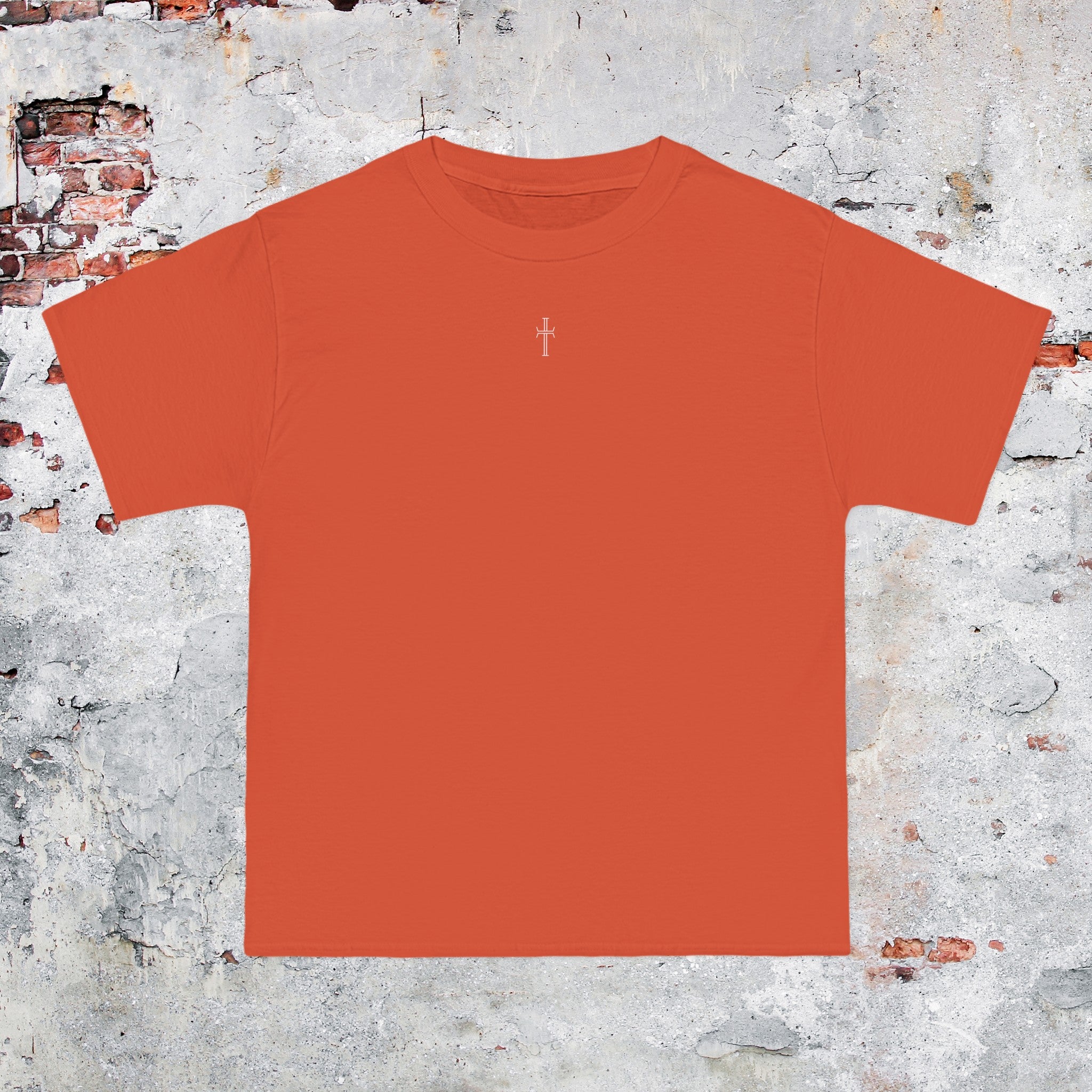 An orange crewneck tee shirt displays the front of the garment as it lays against a brick-patterned wall. A small white Purpose cross logo is printed at the top center. 