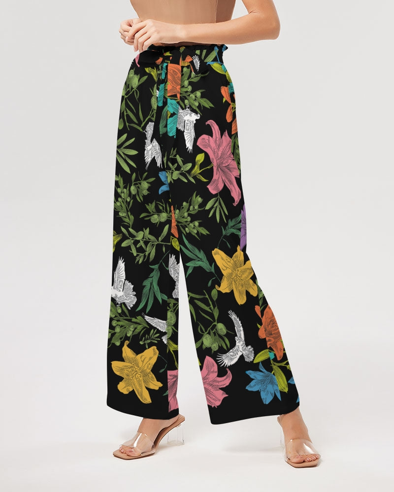 Slightly angled view of bottom half of female model wearing high-waisted, wide-leg black pants with a bold, multi-colored pattern of lilies, white doves, and olive branches in a large, all-over print with self-tie belt and elastic waist.