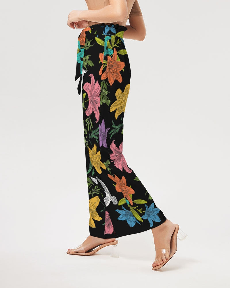 Left view of bottom half of slender woman wearing high-waisted, wide-leg black pants with a bold, multi-colored pattern of lilies, white doves, and olive branches in a large, all-over print with self-tie belt and elastic waist.