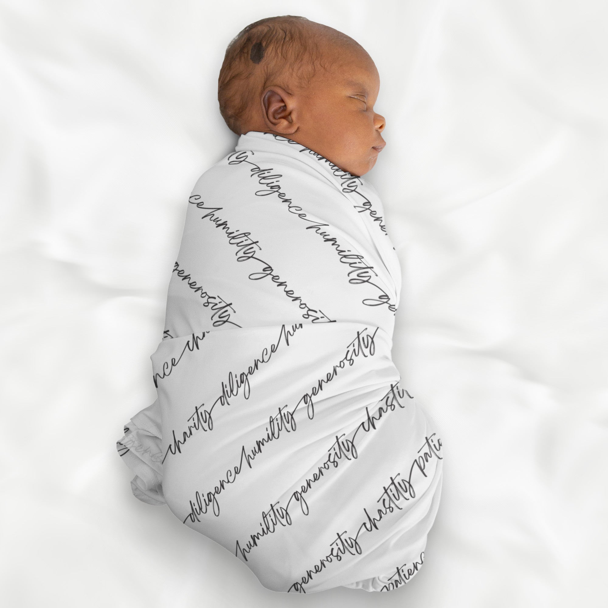 A baby wrapped in a white swaddle blanket with an all-over print of biblical virtues written in black script in continuous strips across the blanket face.