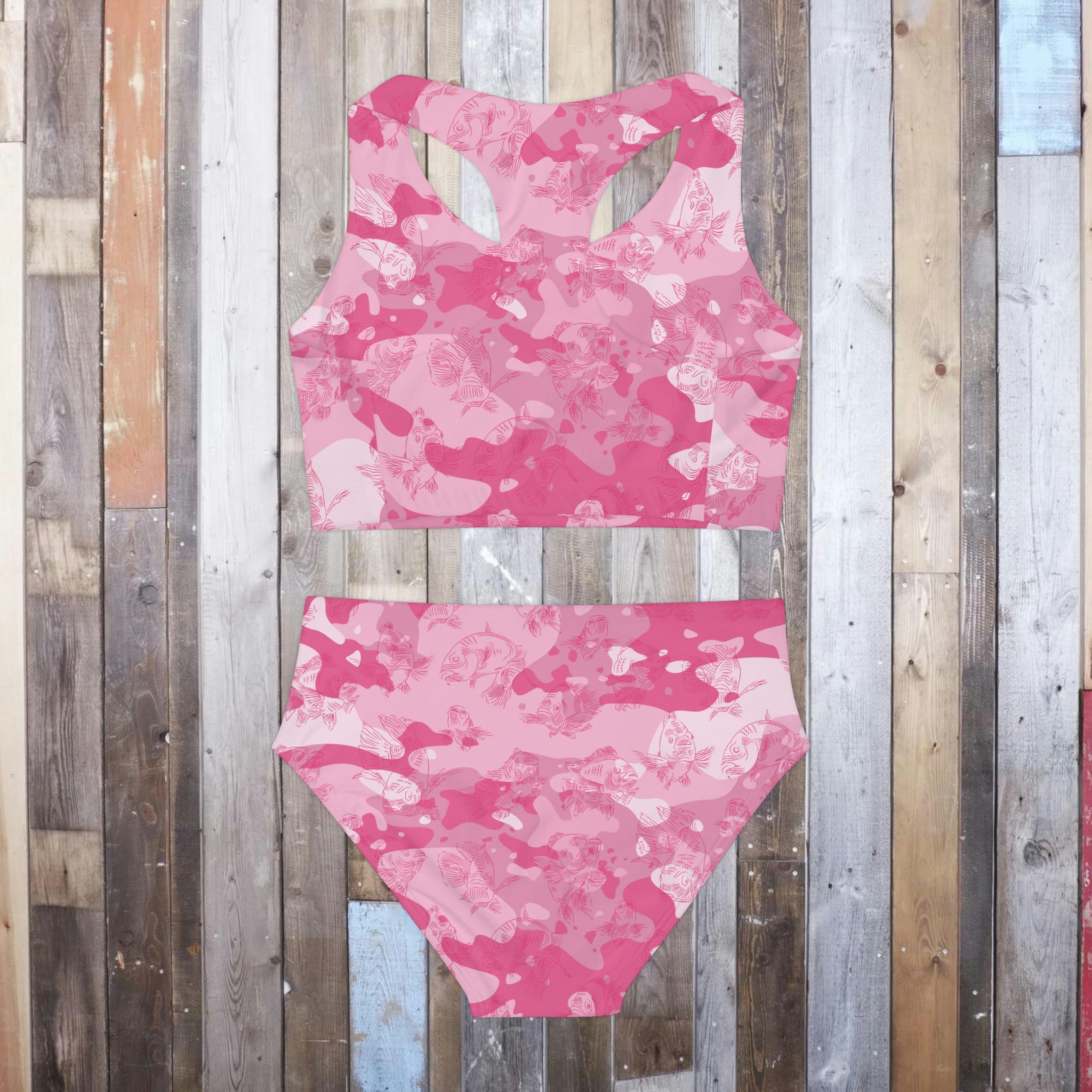 Back view of a two-piece tank-style swimsuit laying flat on a hardwood floor, in a pink-camo design with an all-over swimming carp pattern in dark pink. 