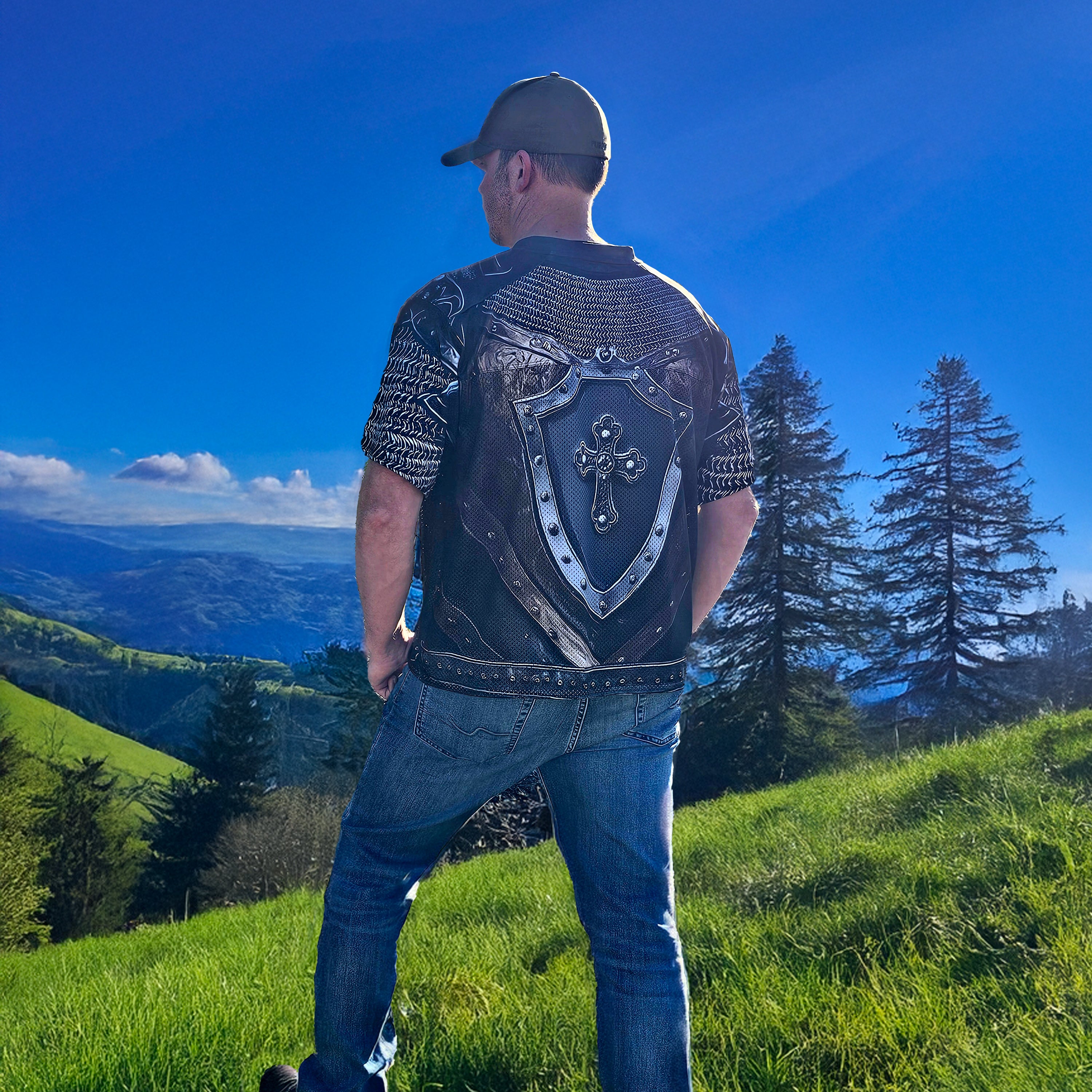 Man wearing a black graphic football jersey and jeans standing on a grassy hill with a scenic view of mountains and trees.