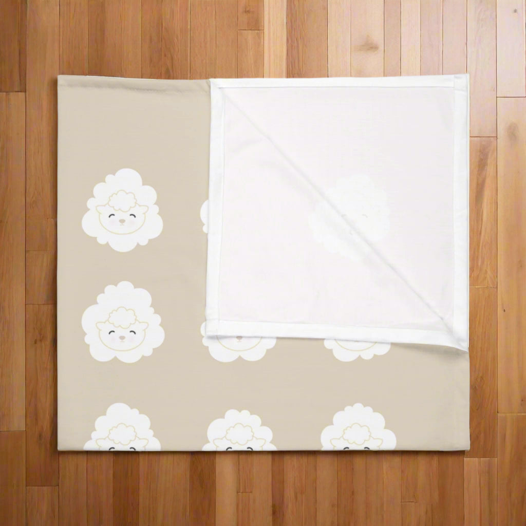 Close-up view of folded beige swaddle blanket with an all-over, white, lamb-face pattern in even rows. Also displays the white opposite side of the blanket.