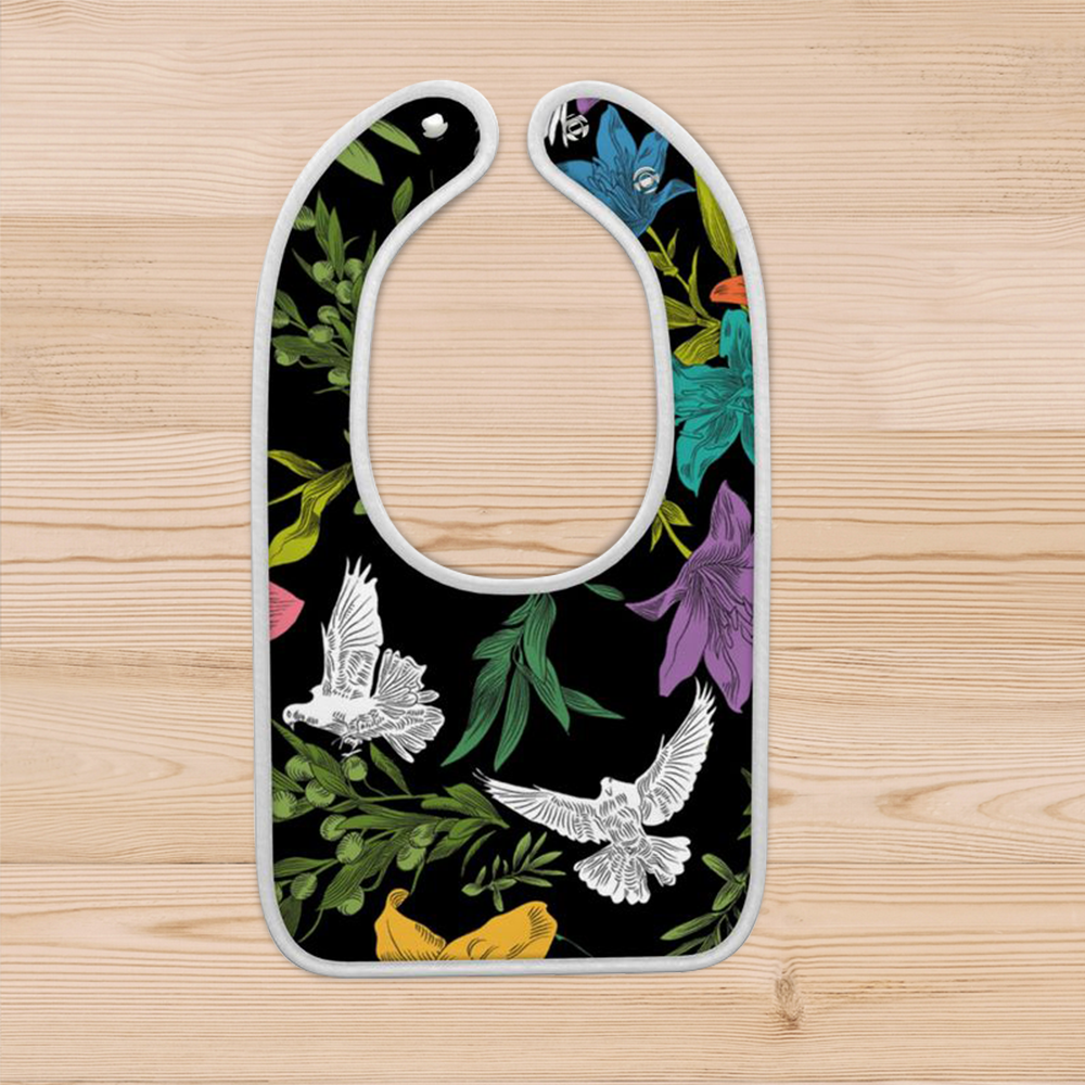 A black baby bib in a multi-colored, all-over print with doves, flowers, and olive branches. It has a white rolled border and two plastic snaps for alternate sizing options.