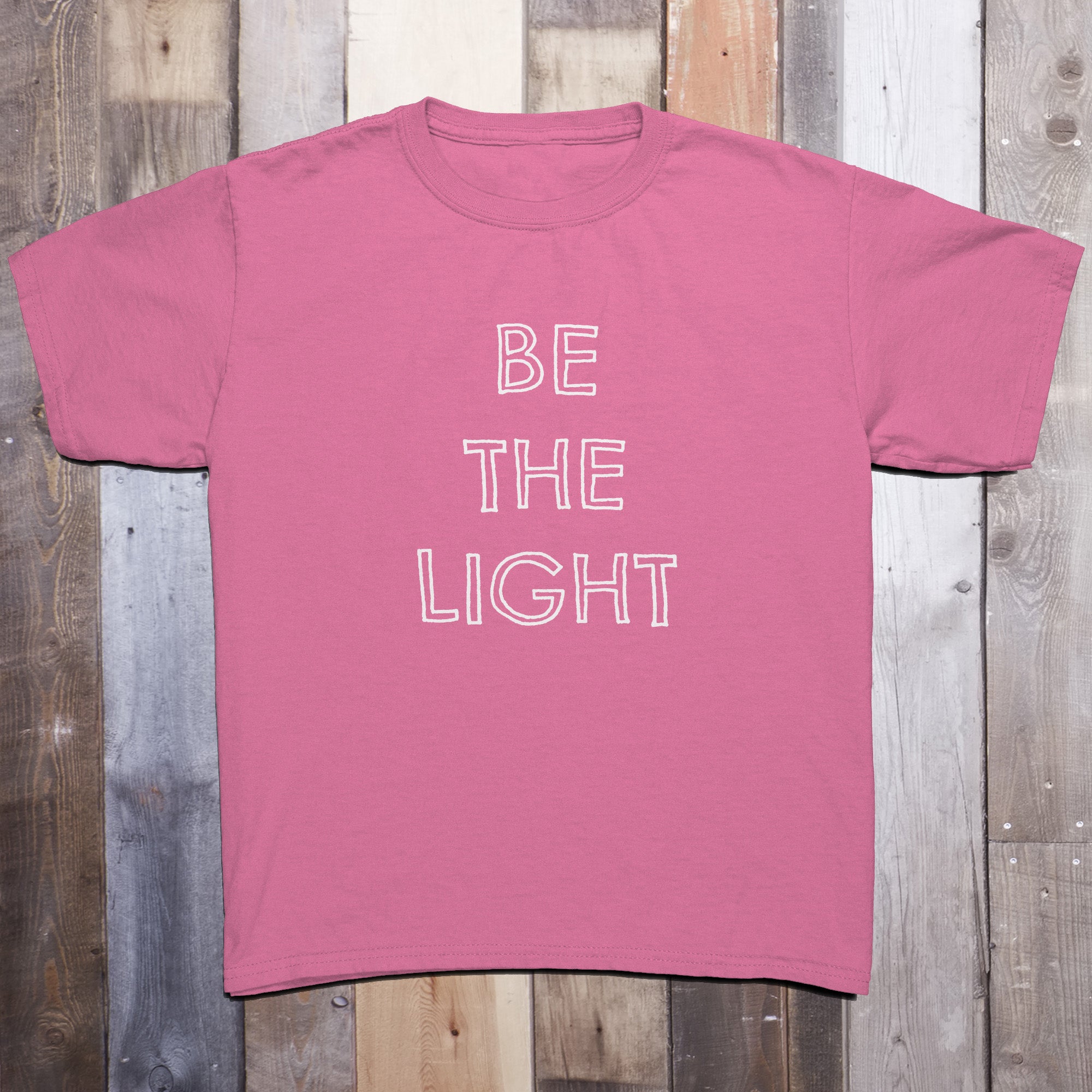 Pink t-shirt with the phrase BE THE LIGHT written in white on the front center of the garment, with each word stacked above the other.