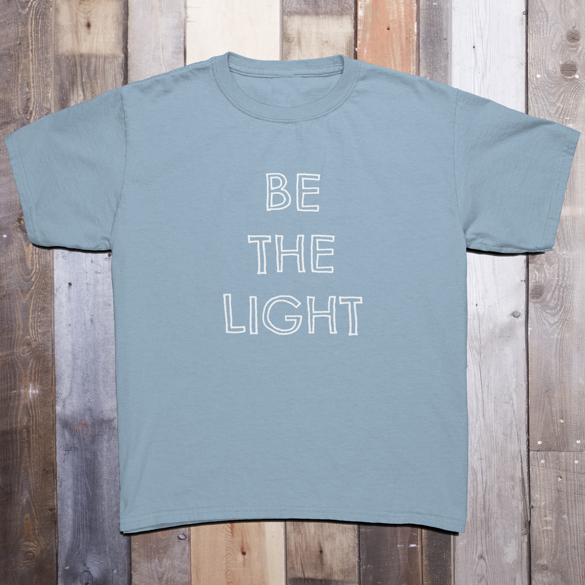 Light blue t-shirt with the phrase BE THE LIGHT written in white on the front center of the garment, with each word stacked above the other.