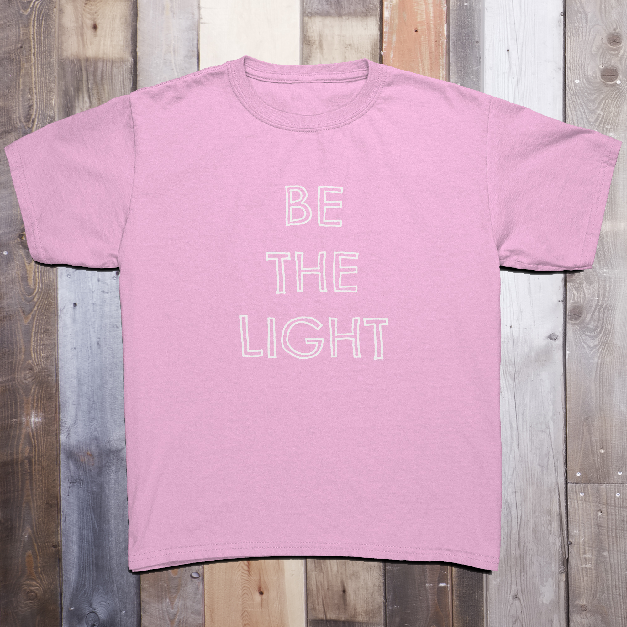 Light pink t-shirt with the phrase BE THE LIGHT written in white on the front center of the garment, with each word stacked above the other.