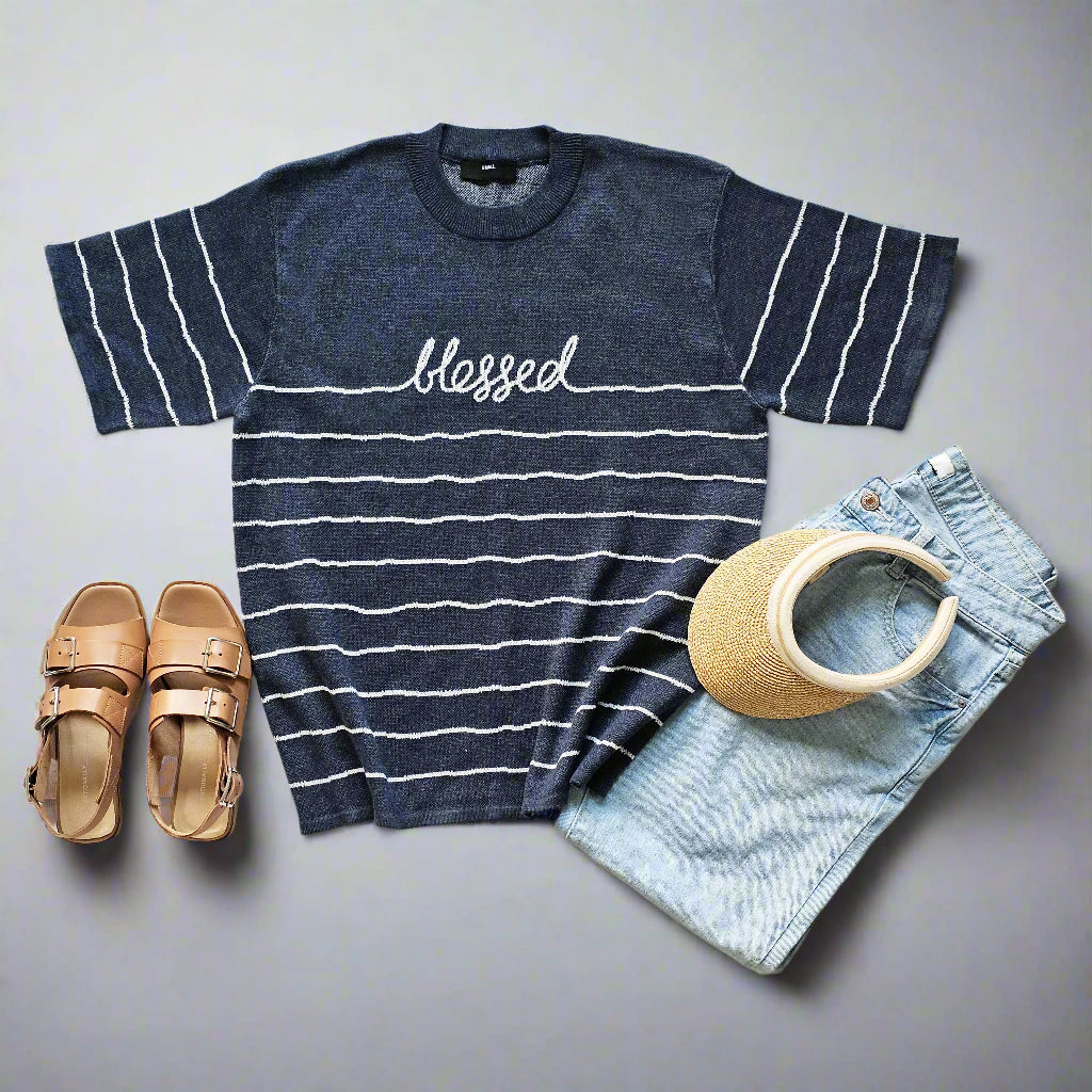 Navy striped t-shirt with 'blessed' text, sandals, visor, and denim shorts on a light background