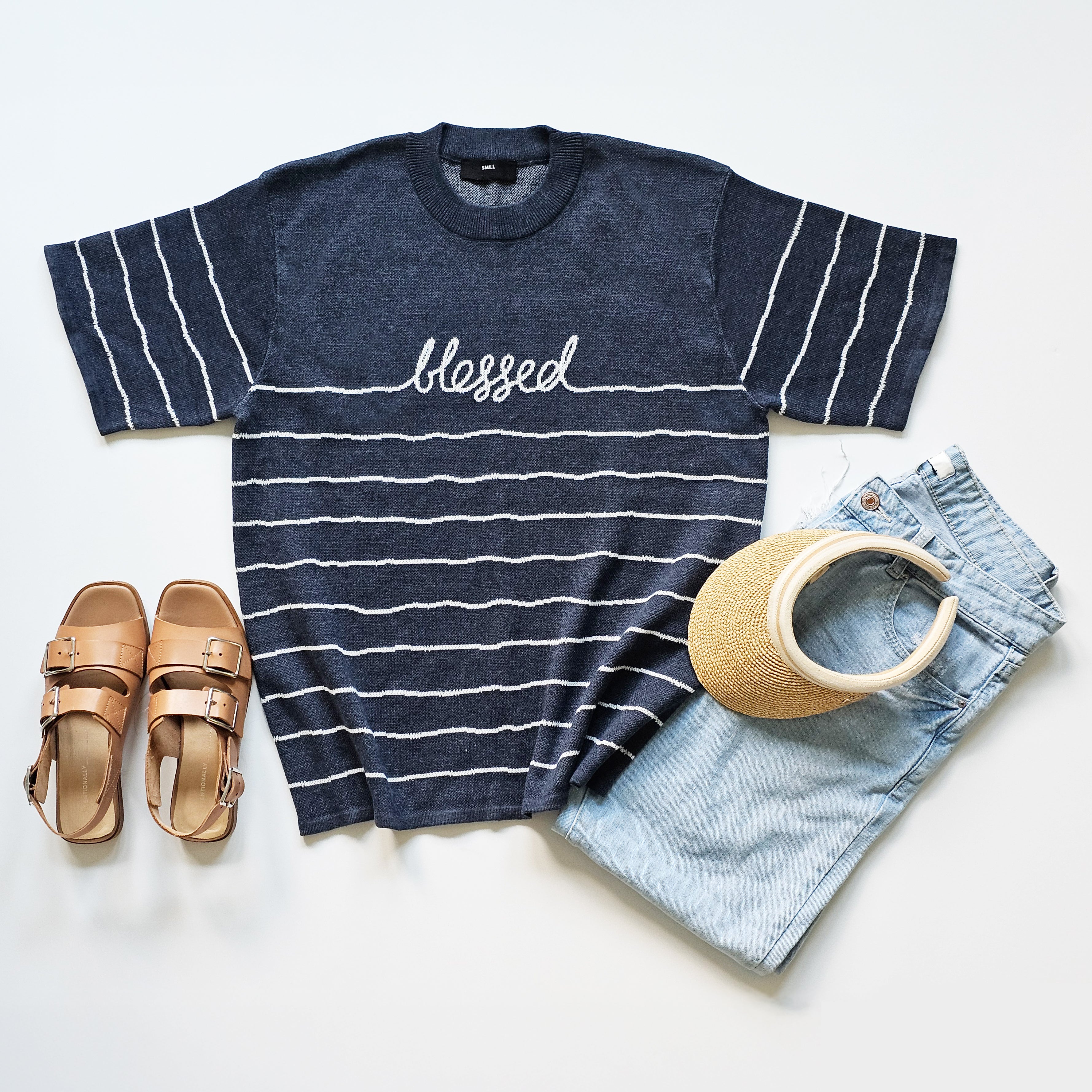 Navy striped t-shirt with 'blessed' text, sandals, visor, and denim shorts on a light background