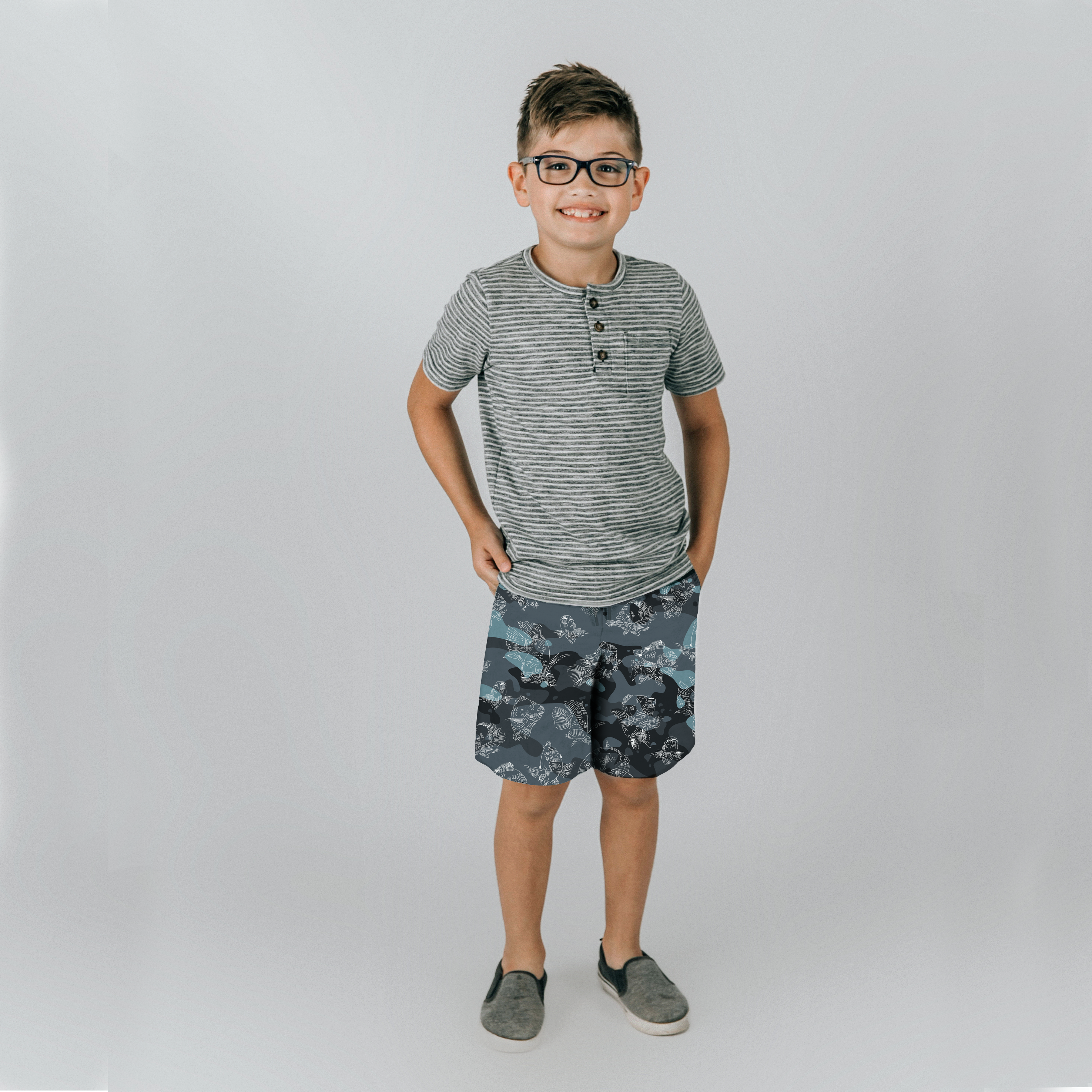 A youth male model in a tee shirt and slip-on shoes, wears blue-camo board shorts with a white all-over swimming carp pattern.