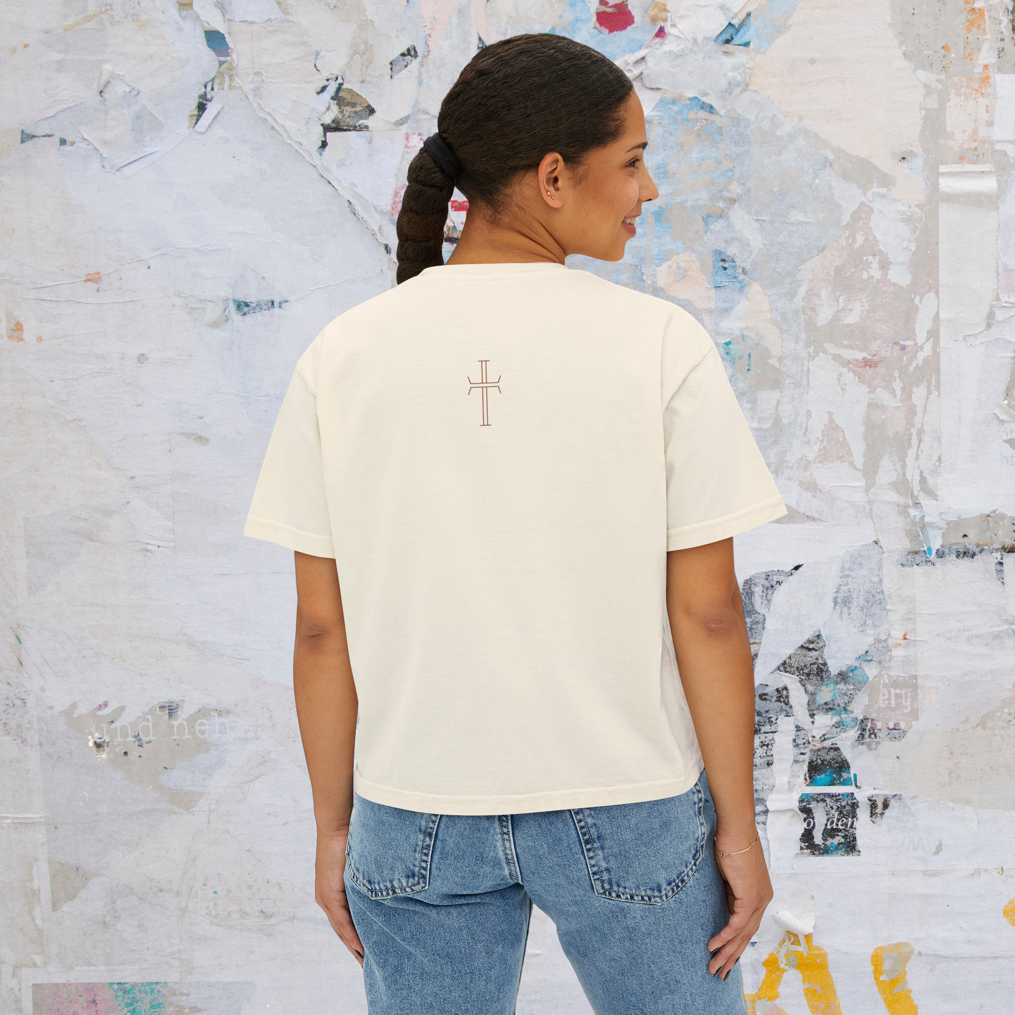 Woman wearing a white t-shirt with a cross design on the back, standing against a textured wall.