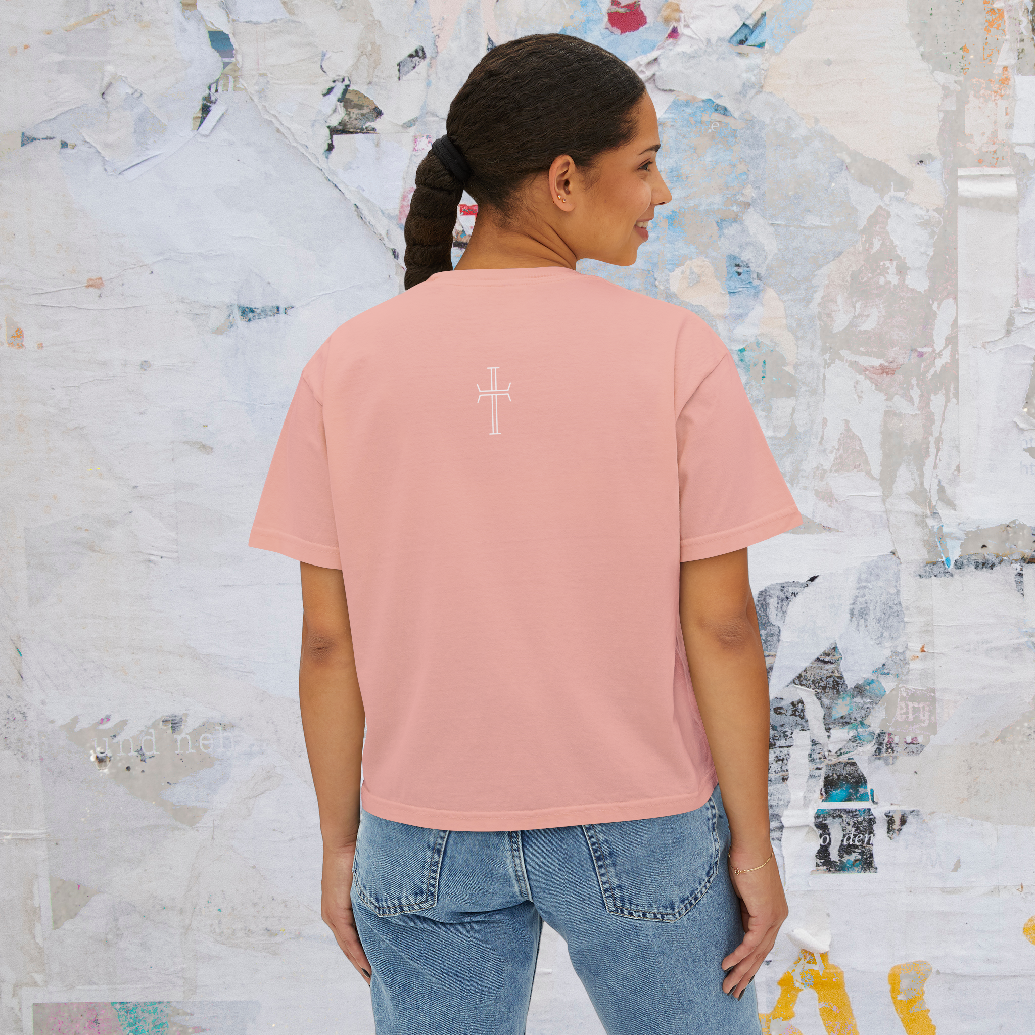 Person wearing a peach colored t-shirt with a cross design on the back, standing against a textured wall.