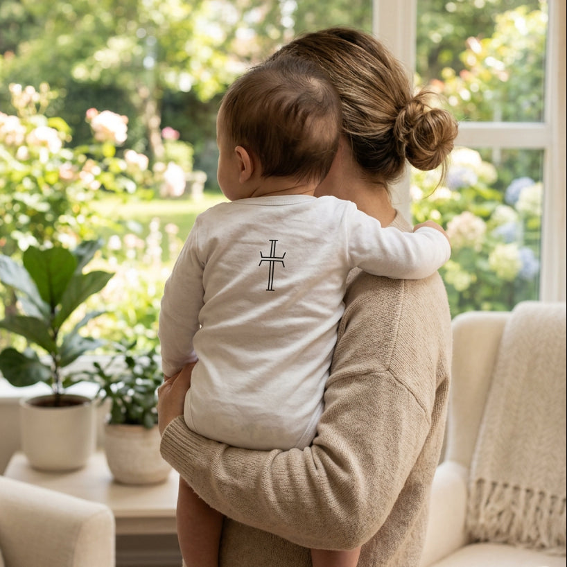 The back side of an infant wearing a white long-sleeve baby bodysuit with a simple black cross graphic printed in the  upper center, displayed flat against a solid white background, being held by his mother in a neutral colored living room next to a window overlooking a lush summer garden.