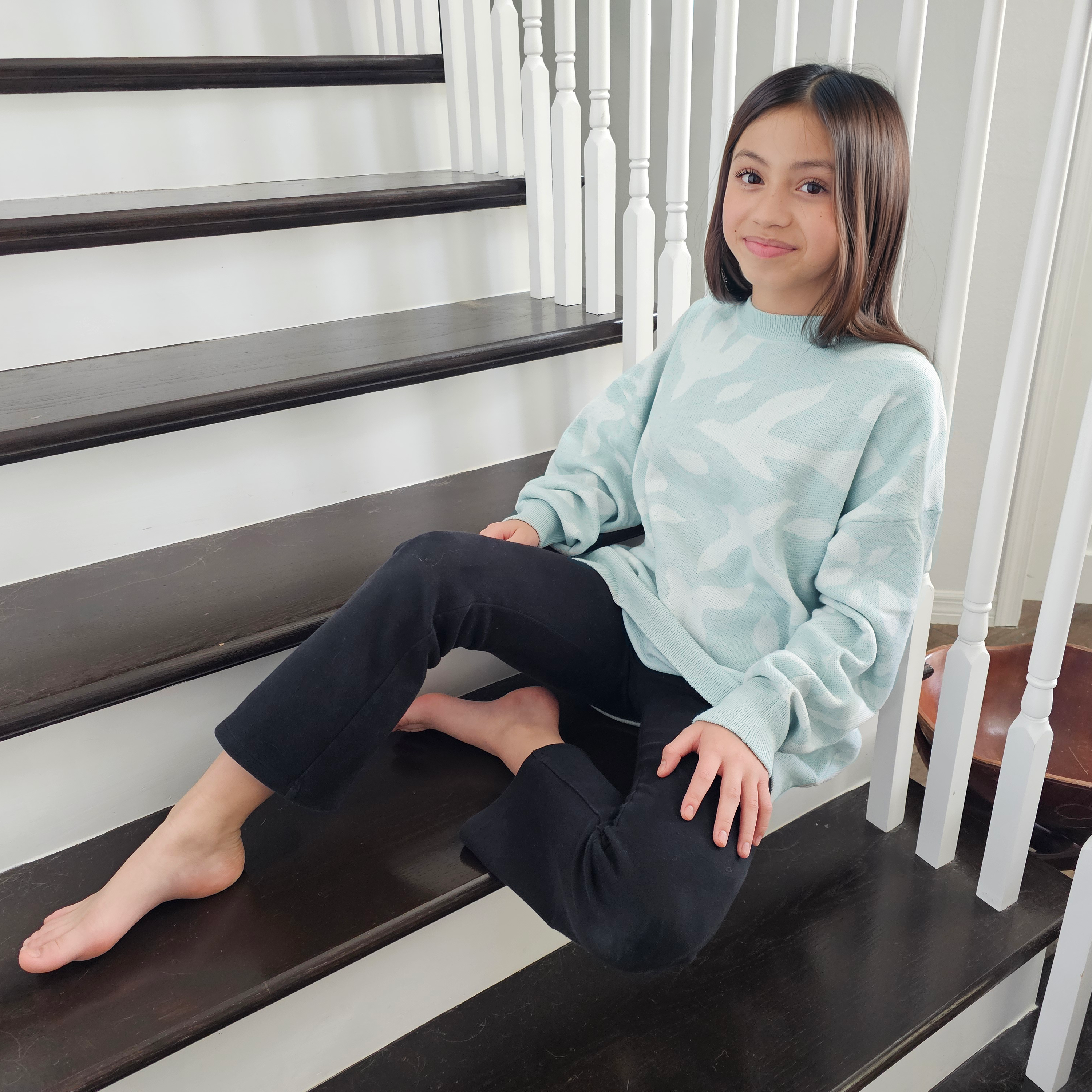 Young girl sitting on a staircase wearing a light blue sweater with white bid and leaf pattern and dark pants.