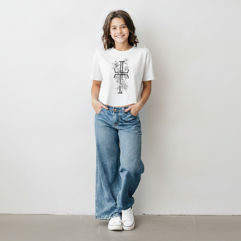 Front of teen girl wearing a cropped white t-shirt with a black and white stylized cross decorated with olive branches and florals, wide-leg blue jeans and white sneakers against a pale wall and gray tiled flooring.