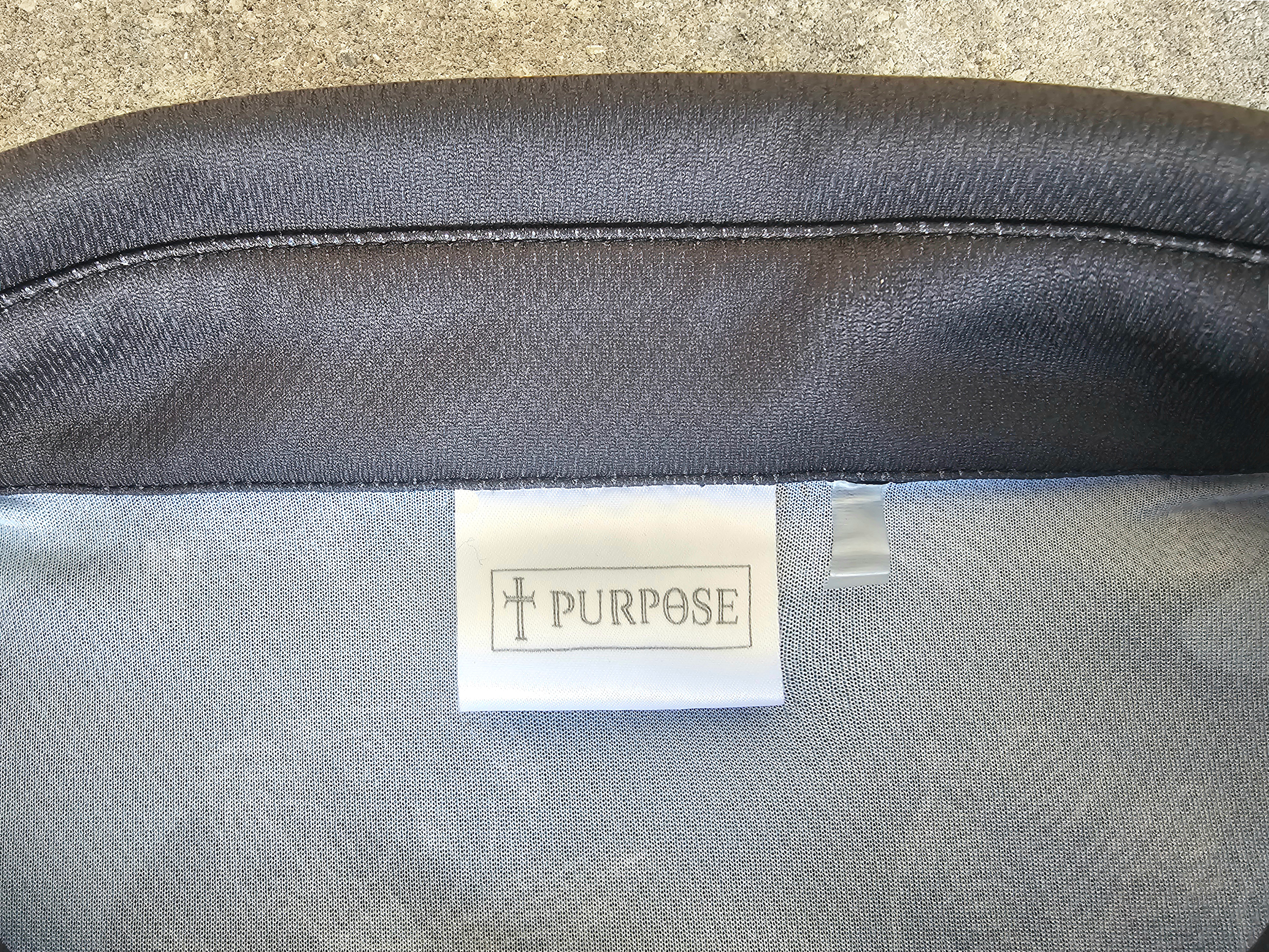 Close-up of a dark gray textured shirt collar with a white fabric label reading 'PURPOSE' and a small cross symbol, set against a neutral gray background.