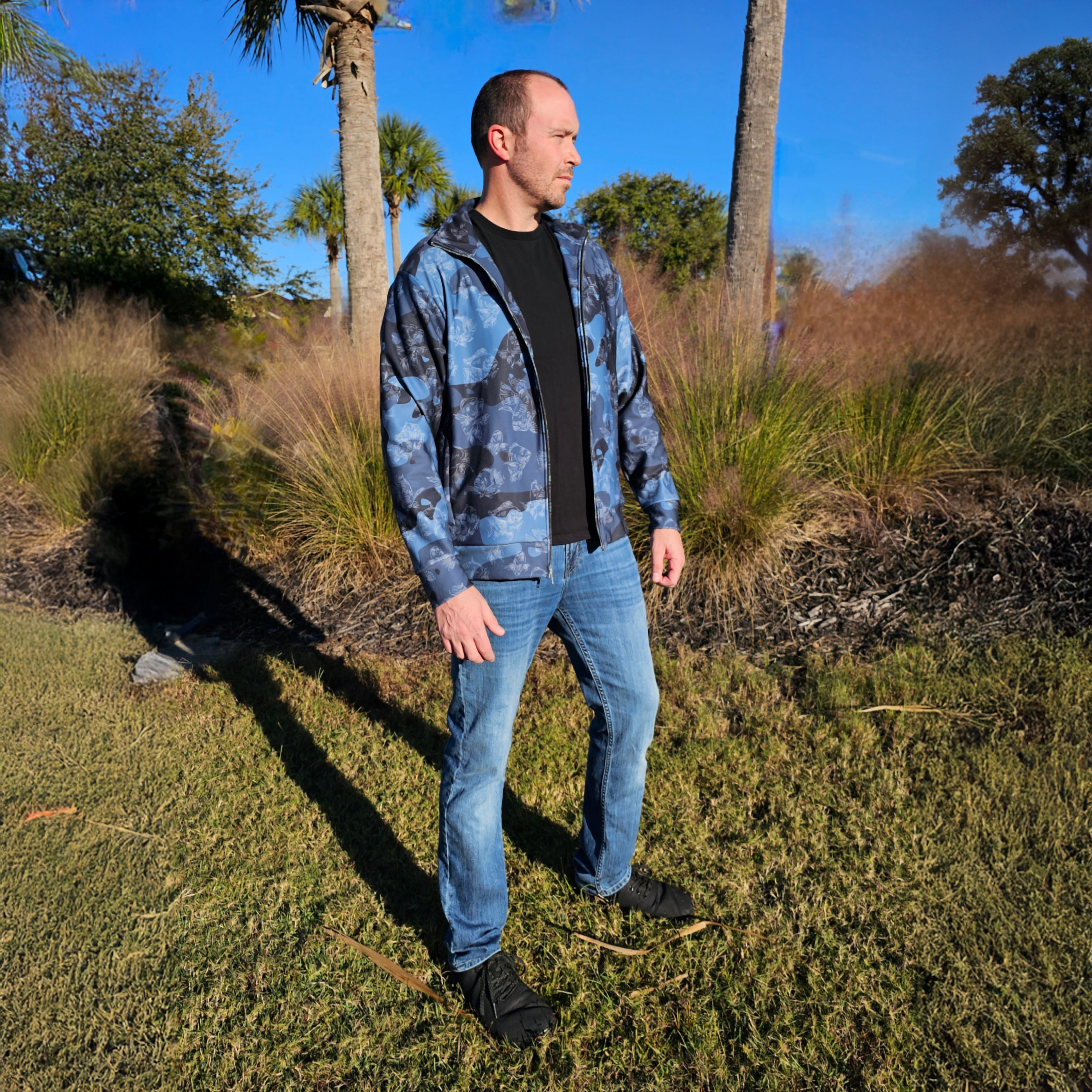 Man wearing a blue camouflage jacket and jeans standing in a grassy area with palm trees.