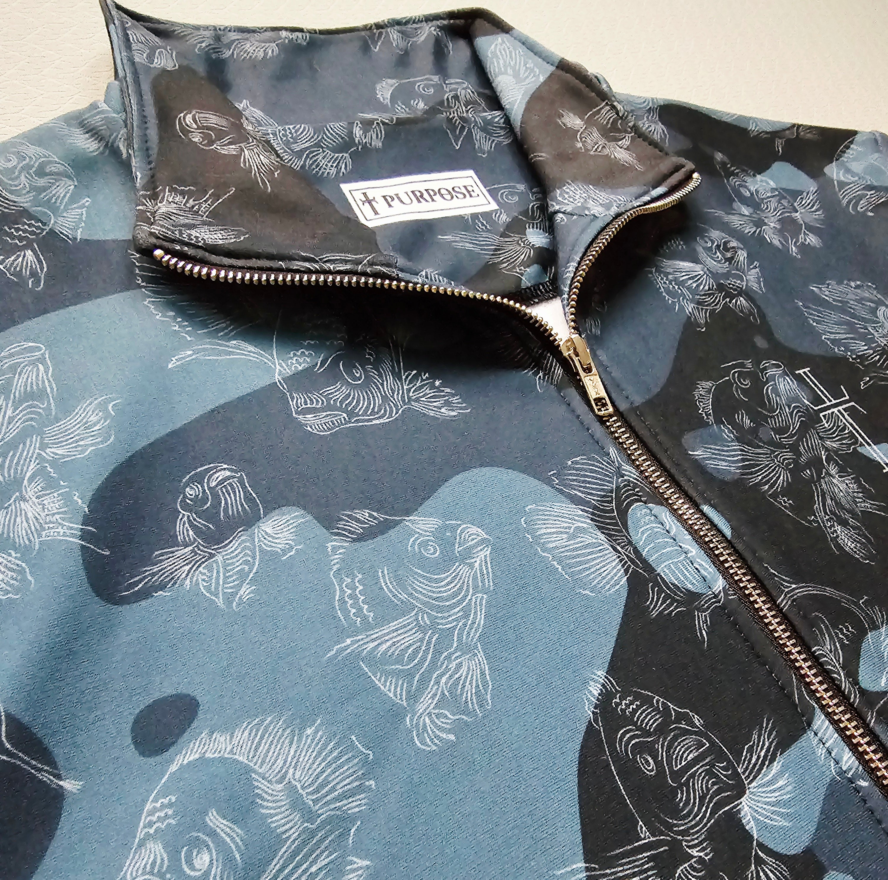 Close-up of blue camouflage jacket with swimming carp pattern and brand label on a neutral background