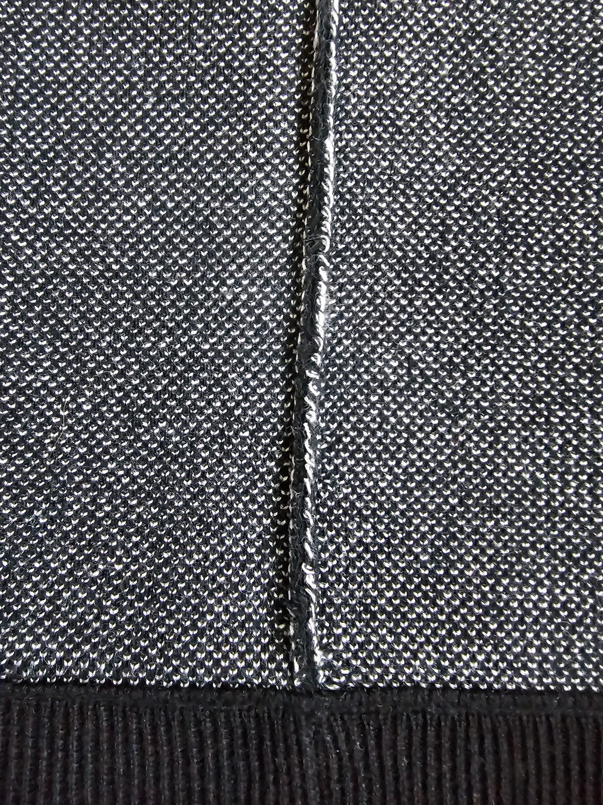 Close-up of a textured knit fabric in a heathered gray and black colorway, showing a central seam and a ribbed black hem.