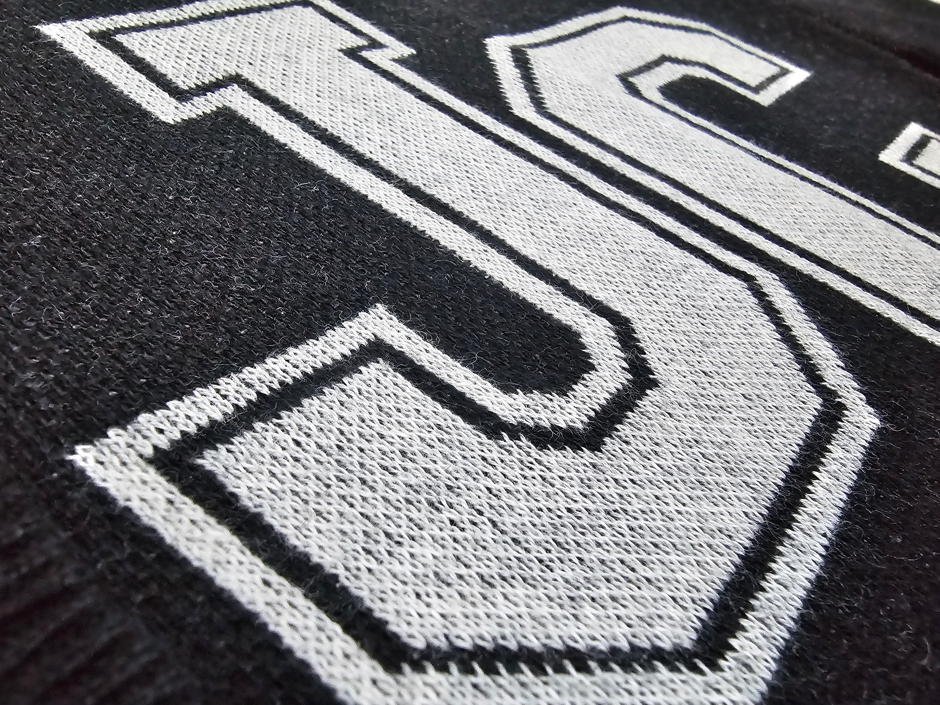Close-up of a black knit fabric with a large white outlined letters 'JC'' knitted across it.