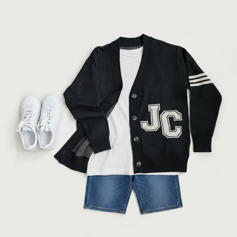 A styled flat-lay outfit featuring a black varsity cardigan with 'JC' lettering, blue denim shorts, a white t-shirt, and white sneakers, arranged on a neutral gray background.