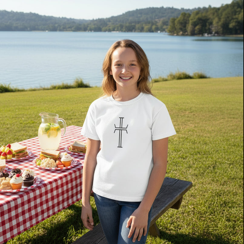Girl wearing a white t-shirt with stylized Christian cross on a white background, in a park next to a placid lake and a picnic table of delicious foods on a red and white checkered table cloth.