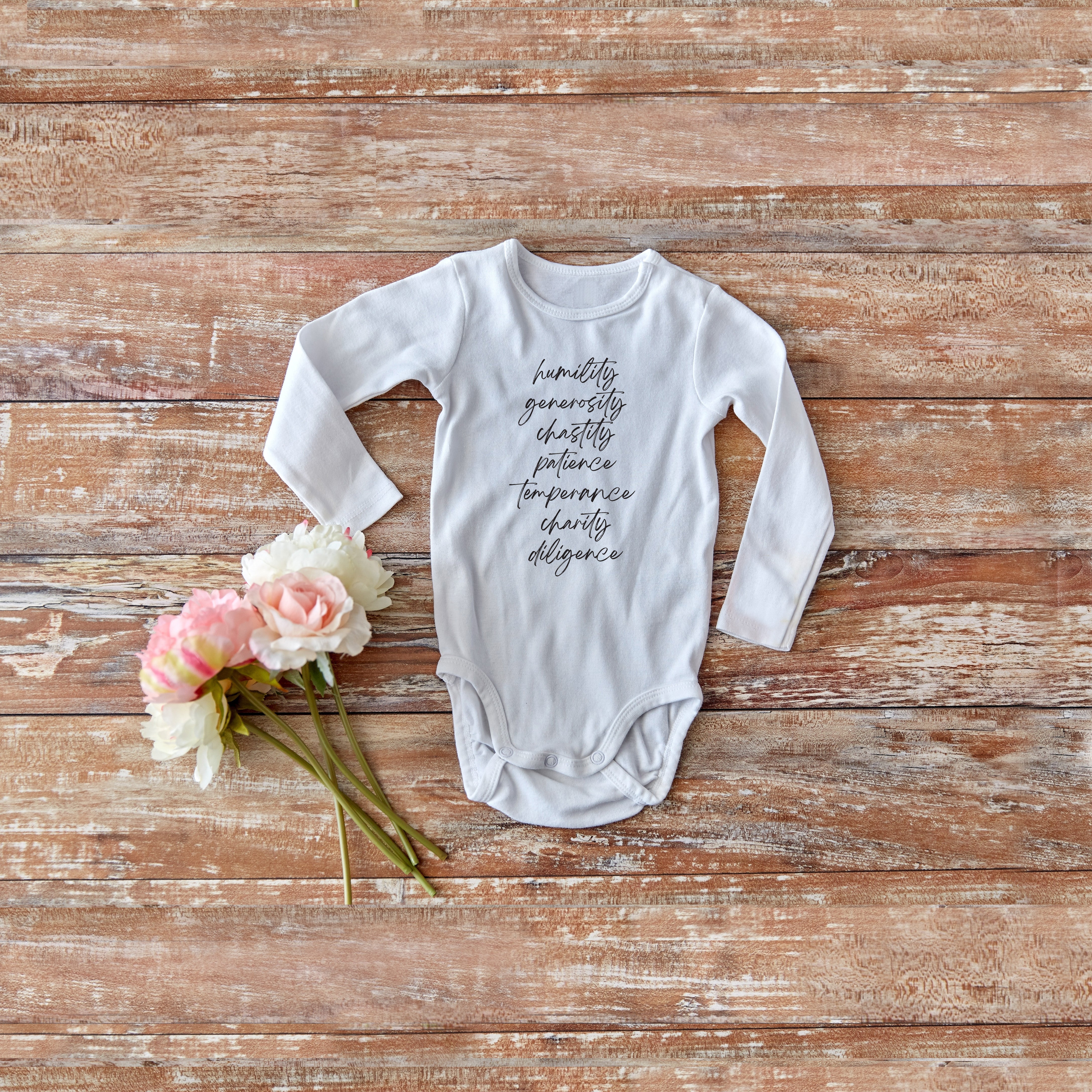 A white baby onsie with seven biblical virtues written in script down the center of the garment, with one word stacked evenly above the other.