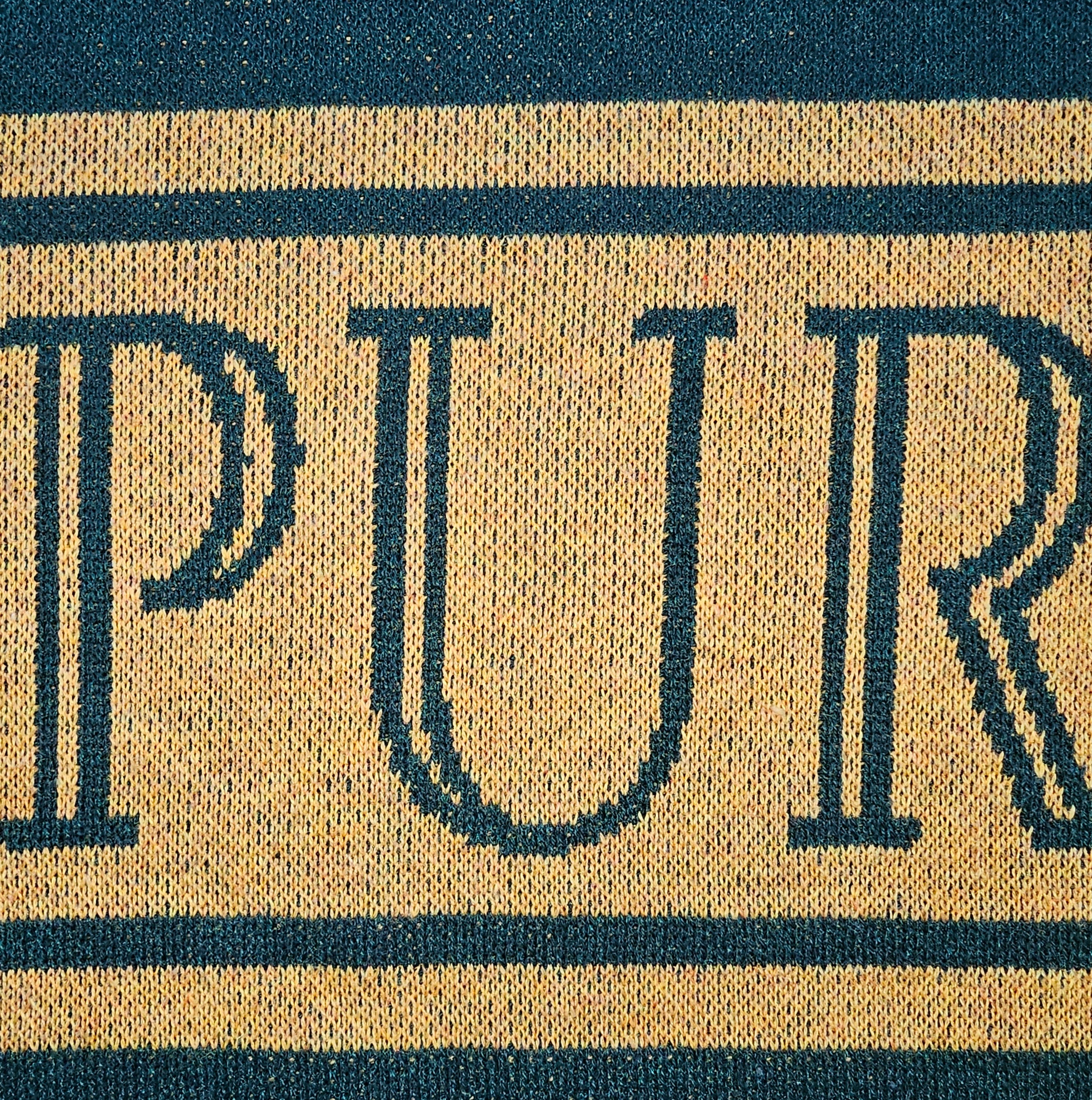 A close up of the knitted "PUR" of the Purpose word on the sweater. The dual-colored jacquard knit is detailed.