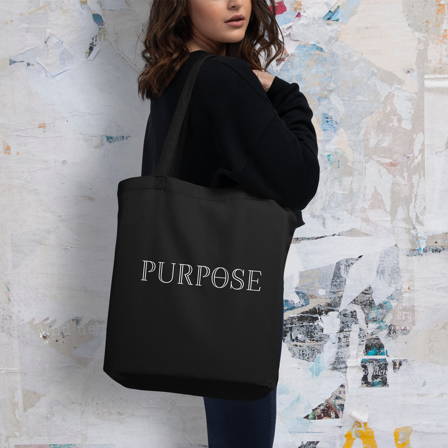 A female model displays the back of a black tote bag over her shoulder. It has two long handles and the white Purpose worded logo printed on the back of the bag.