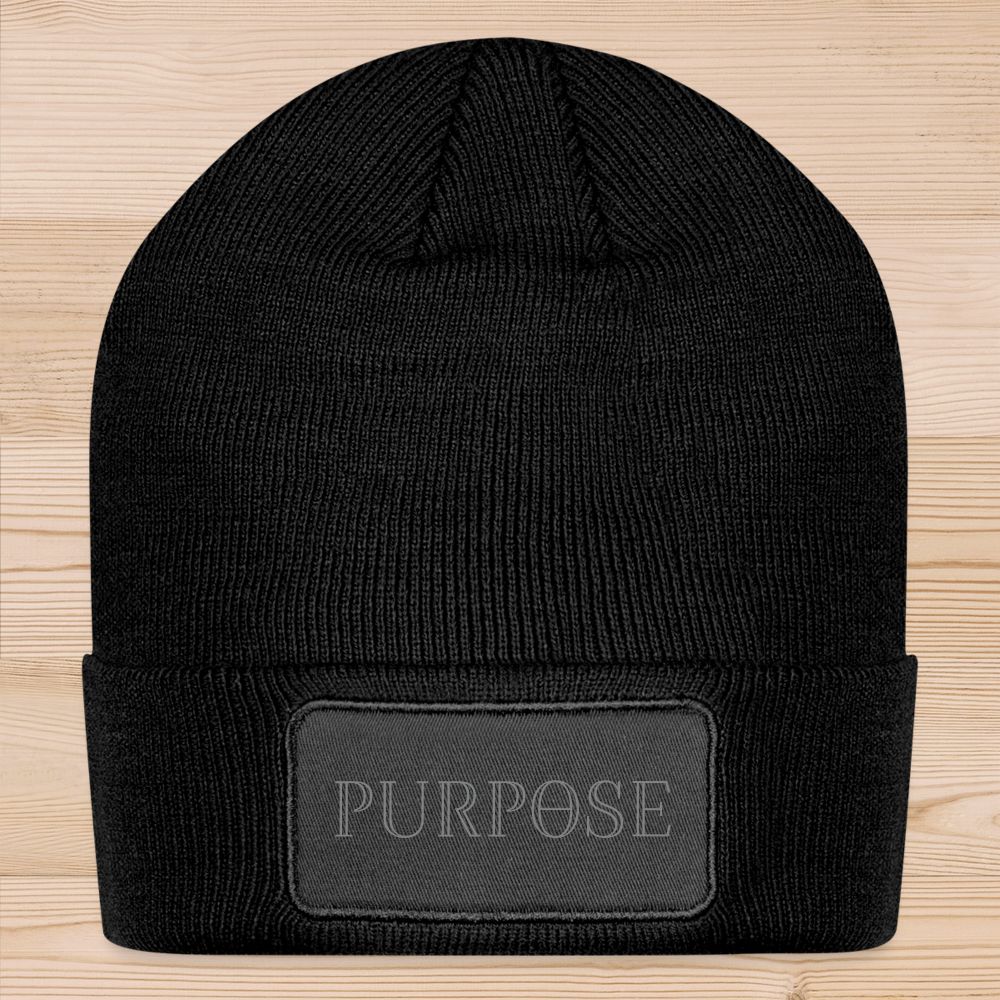 A black knit beanie  laying flat, with a twill, rectangular patch with rounded corners sewn to the front folded brim, printed with the dark gray Purpose worded logo.