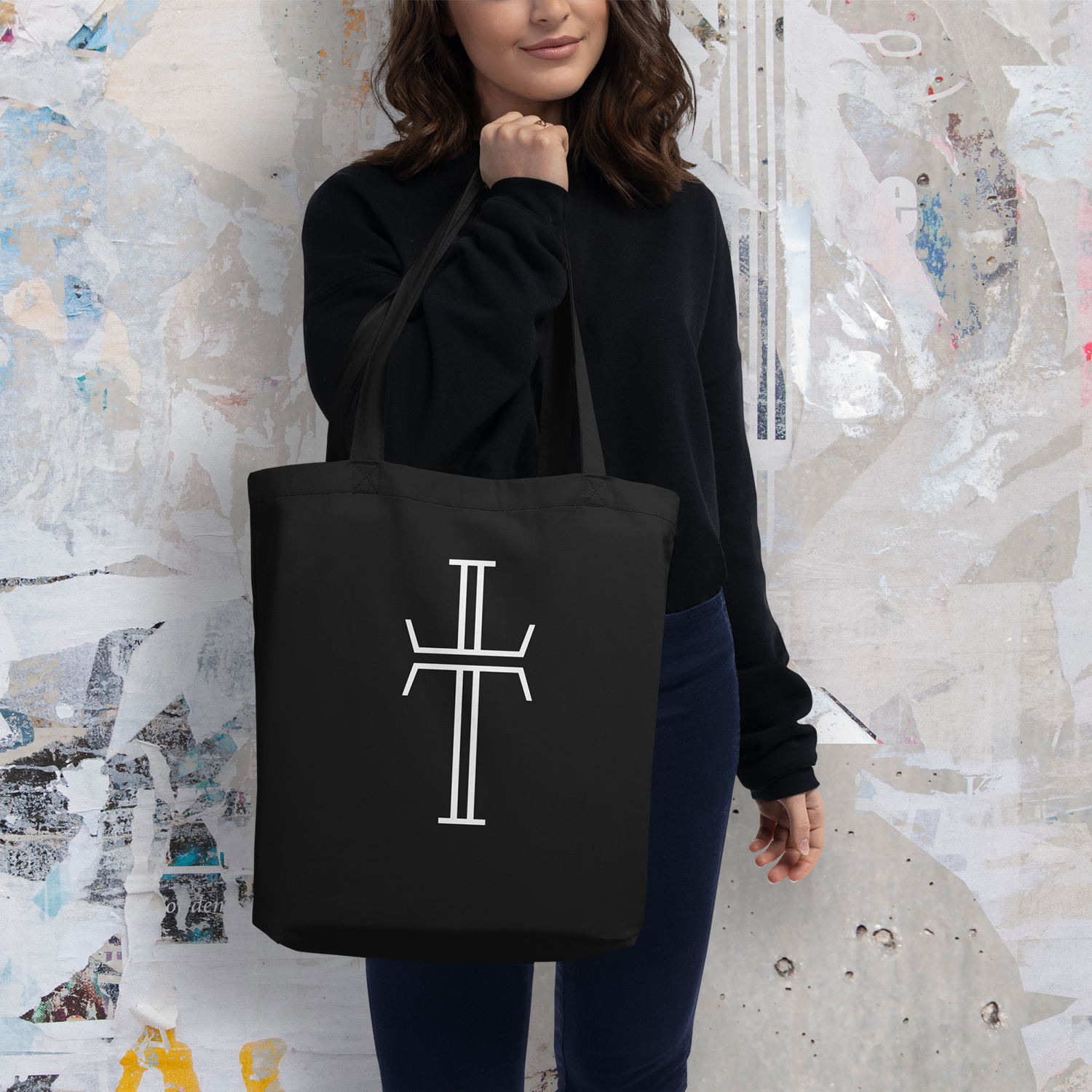 A female model holds a black tote bag with a large white Purpose cross logo. The bag has long double handles.