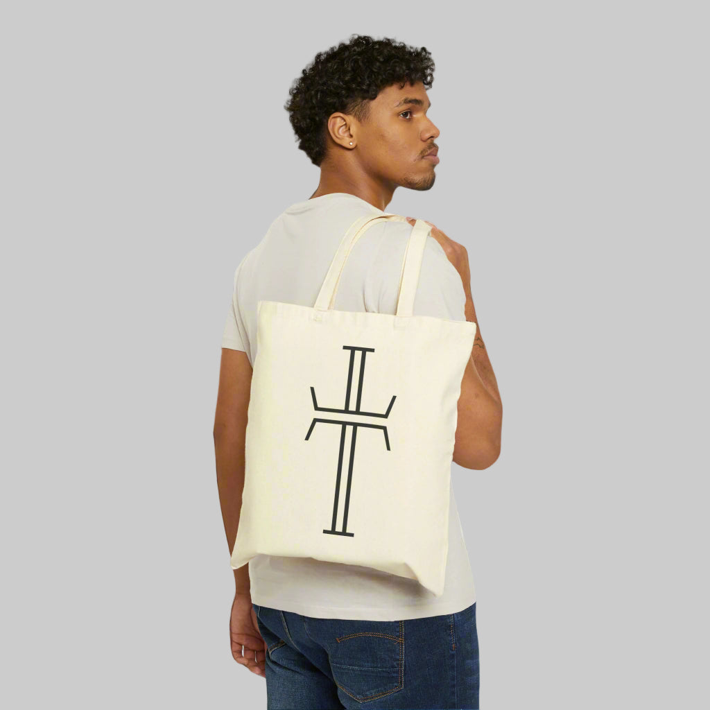 A male model in a tee shirt and jeans is slinging a tote bag with long double handles over the back of his shoulder. The natural canvas bag has a black Purpose cross logo on the front.