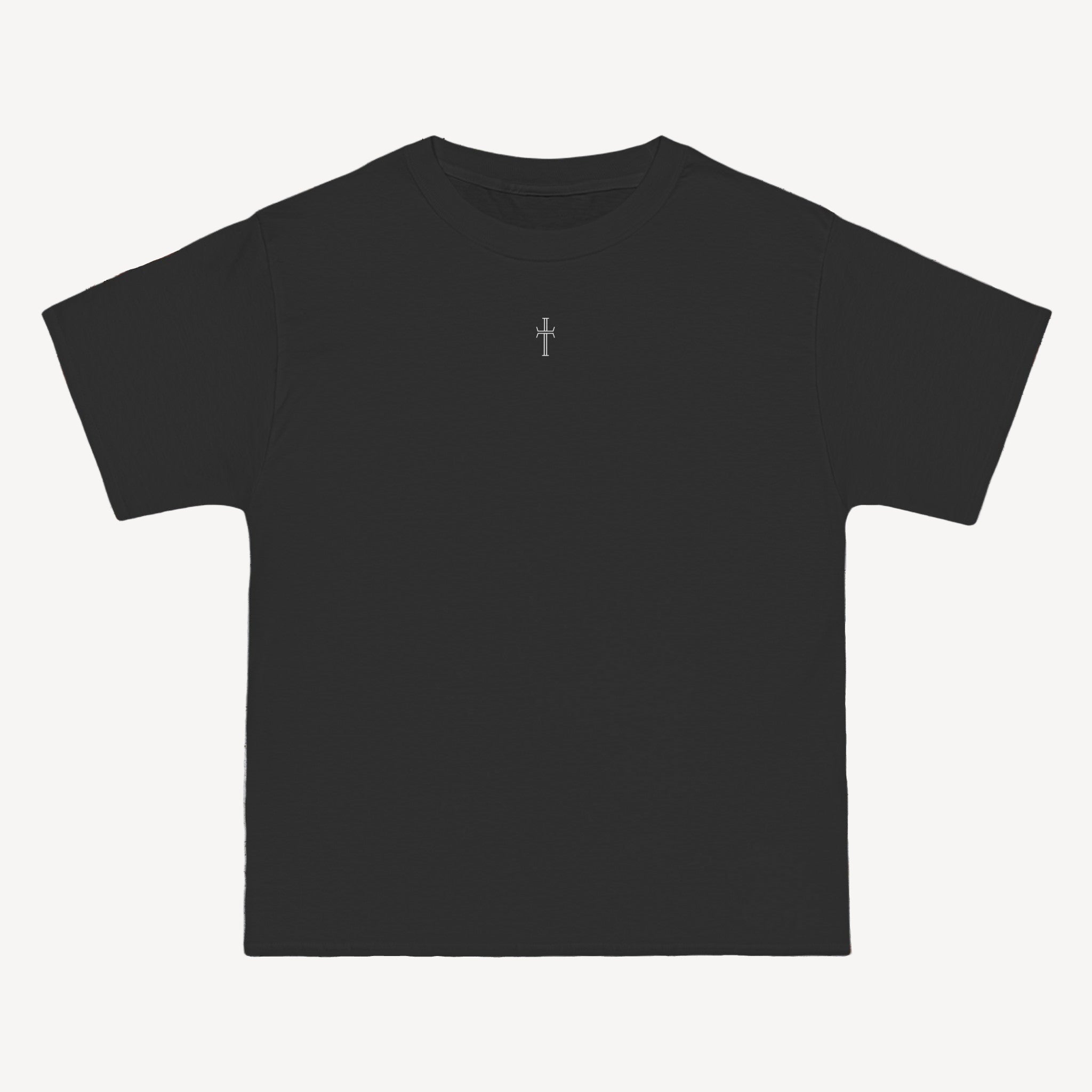 A black crewneck tee shirt displays the front of the garment as it lays against a brick-patterned wall. A small white Purpose cross logo is printed at the top center. 