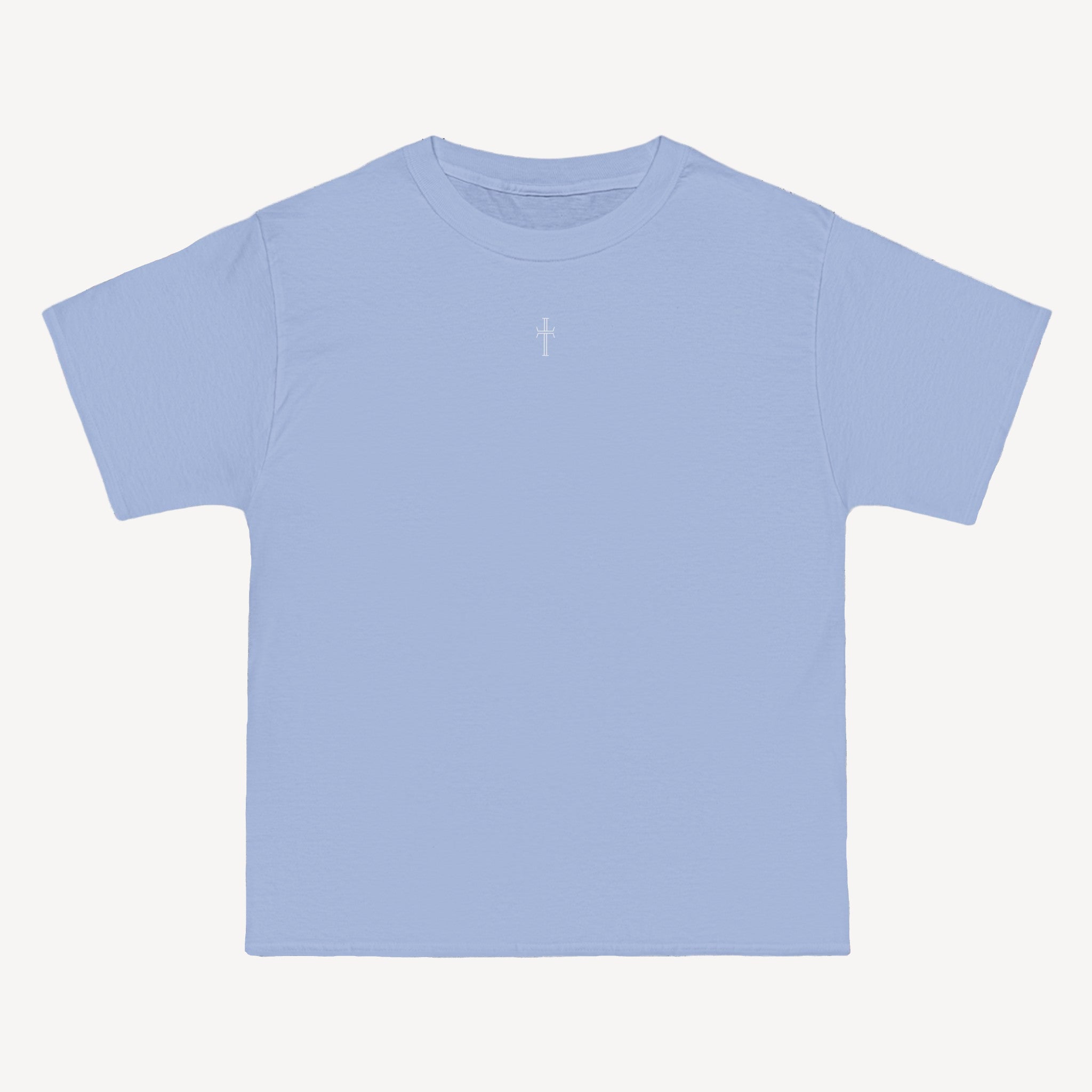 A light blue crewneck tee shirt displays the front of the garment as it lays against a brick-patterned wall. A small white Purpose cross logo is printed at the top center. 