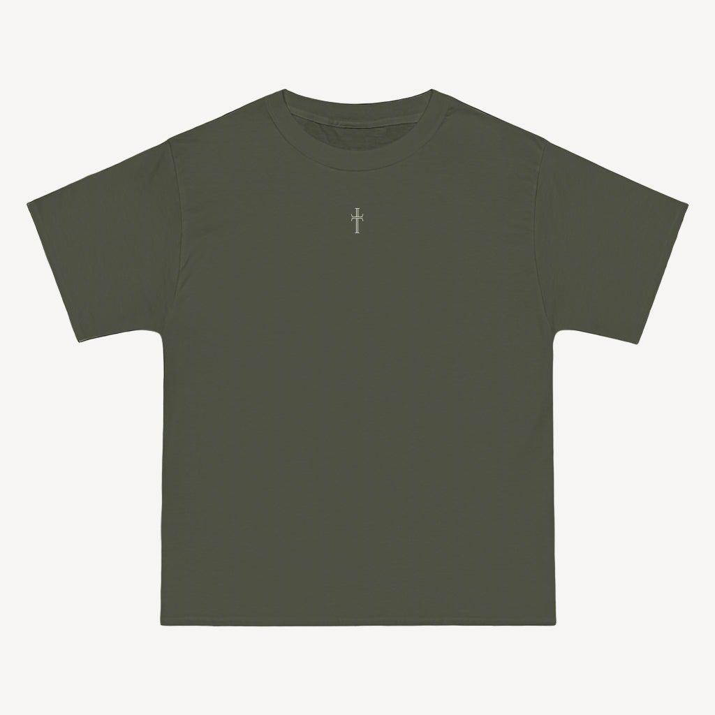 An army green crewneck tee shirt displays the front of the garment as it lays against a brick-patterned wall. A small white Purpose cross logo is printed at the top center. 