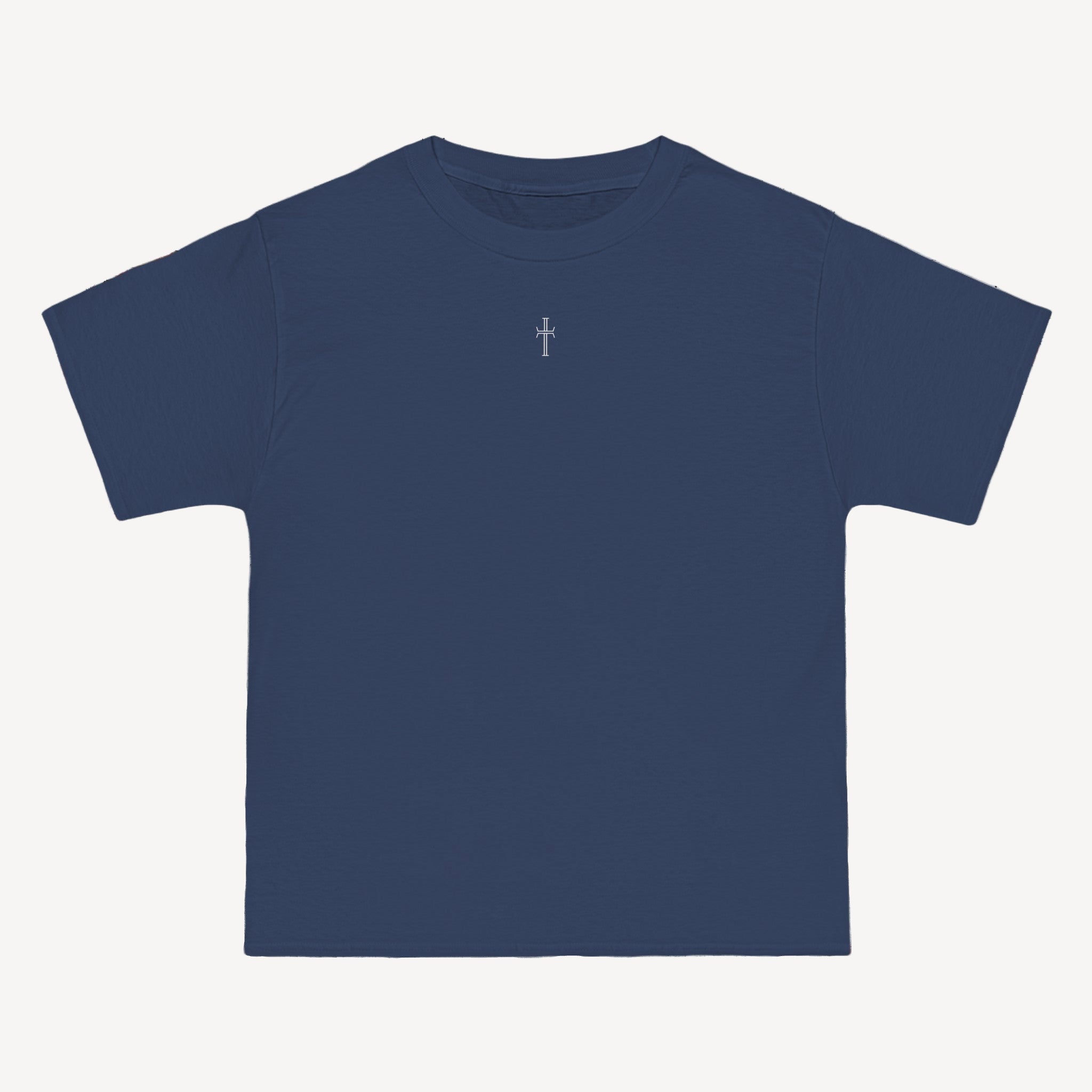 A denim blue crewneck tee shirt displays the front of the garment as it lays against a brick-patterned wall. A small white Purpose cross logo is printed at the top center. 