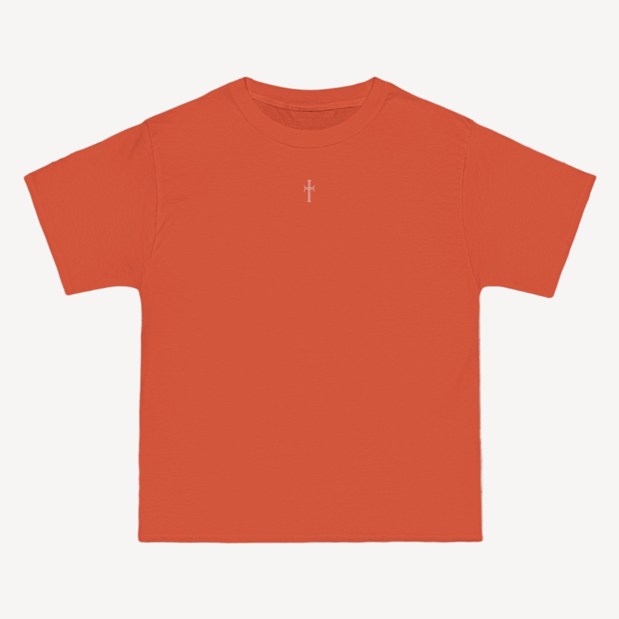 An orange crewneck tee shirt displays the front of the garment as it lays against a brick-patterned wall. A small white Purpose cross logo is printed at the top center. 