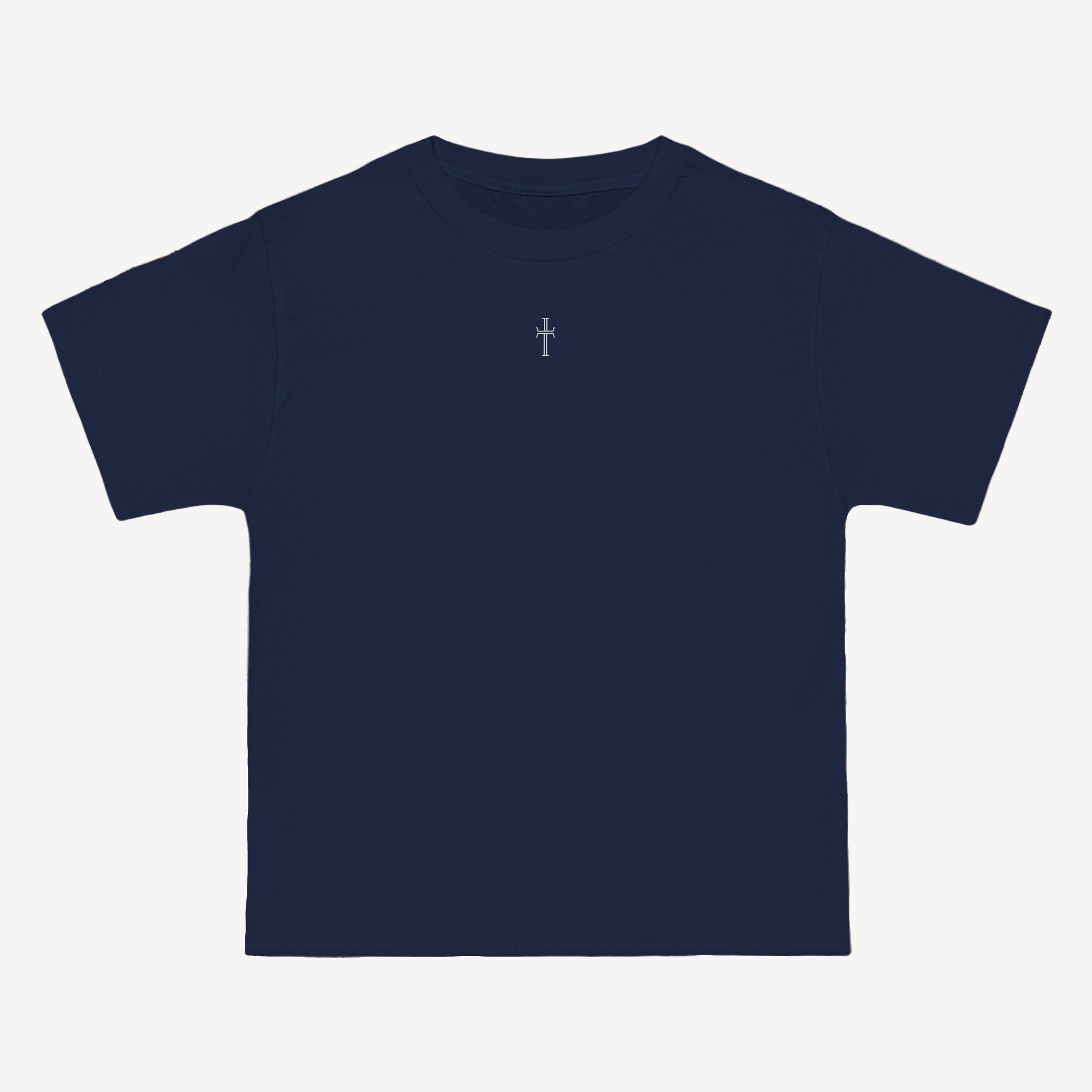 A navy crewneck tee shirt displays the front of the garment as it lays against a brick-patterned wall. A small white Purpose cross logo is printed at the top center. 
