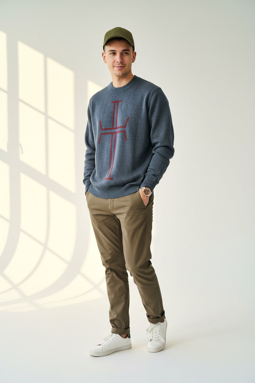 Young man wearing a blue sweater with red logo, olive green cap, brown pants, and white sneakers on a light blue background