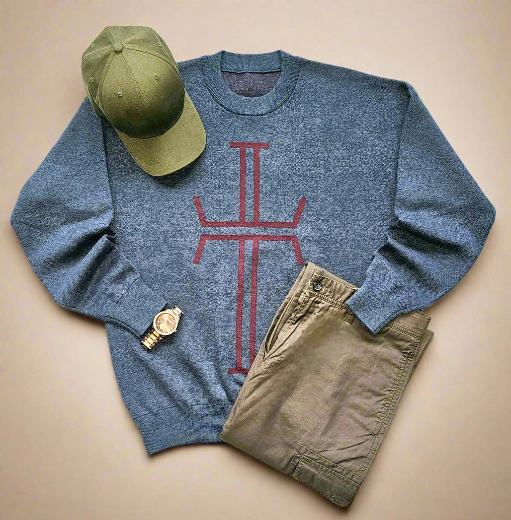 A marled navy blue sweater lays flat against a white background. It has a burgundy stylized cross knitted on the entire front of the sweater. A pair of dark khaki pants, an olive baseball cap, and a wristwatch are arranged around the sweater.