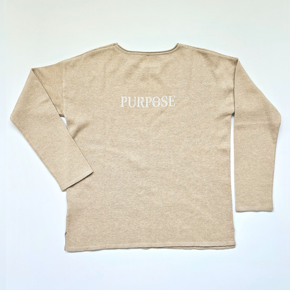 The back of the beige, straight-hemmed sweater. The word "Purpose" is knitted on the upper center back of the sweater in white capital letters.