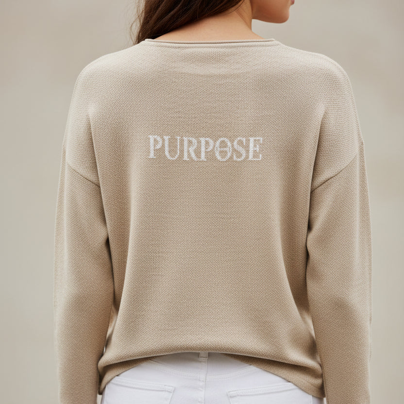 The back of the beige, straight-hemmed sweater. The word "Purpose" is knitted on the upper center back of the sweater in white capital letters.