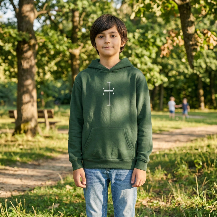 Teen boy wearing a dark green hoodie with a white stylized cross on the center chest, and blue jeans in a park setting.