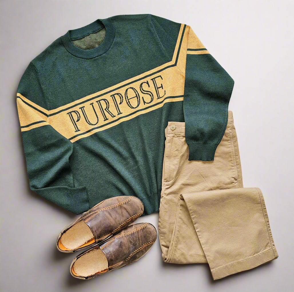 A forest green sweater lays flat against a white background. On the sweater, there is one wide mustard-colored stripe between two thinner stripes of the same color, running along the center of the chest and upper arms. The word "Purpose" is written in forest green in all capital letters on the wide chest stripe. The sweater is arranged next to a pair of khaki pants and a pair of brown loafers.