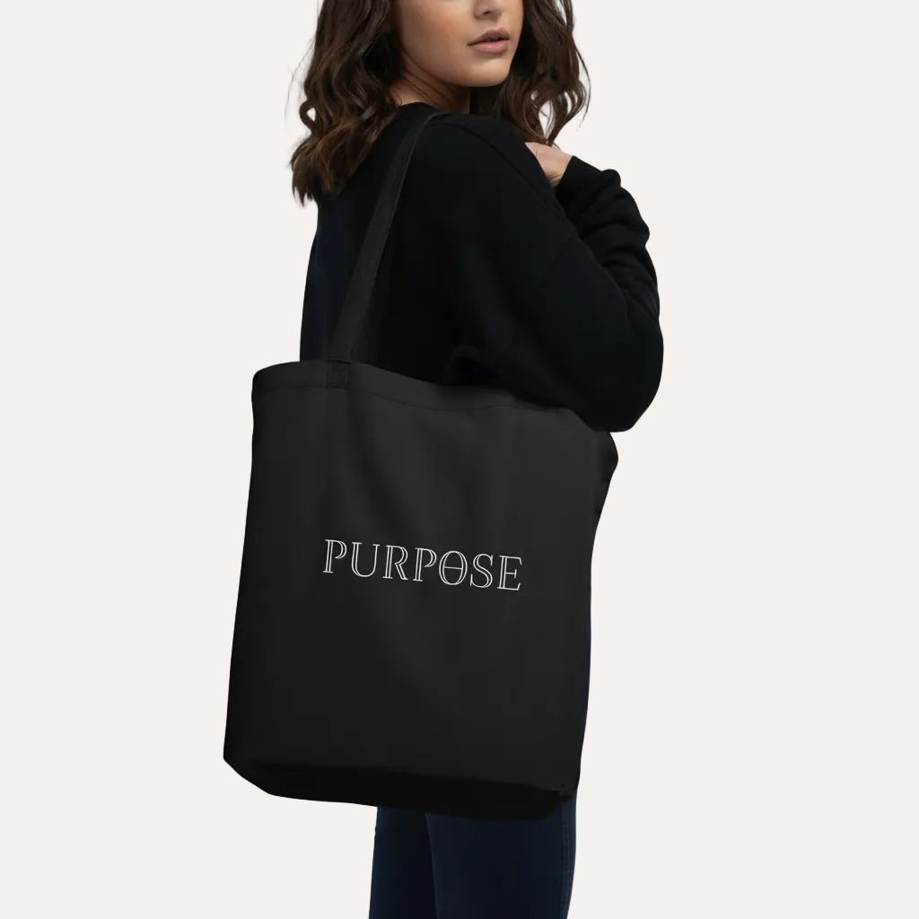 A woman wearing a black sweatshirt and jeans carries a black canvas tote bag over her shoulder. The bag features the word 'PURPOSE' printed in white serif font on the back, set against a plain white background.