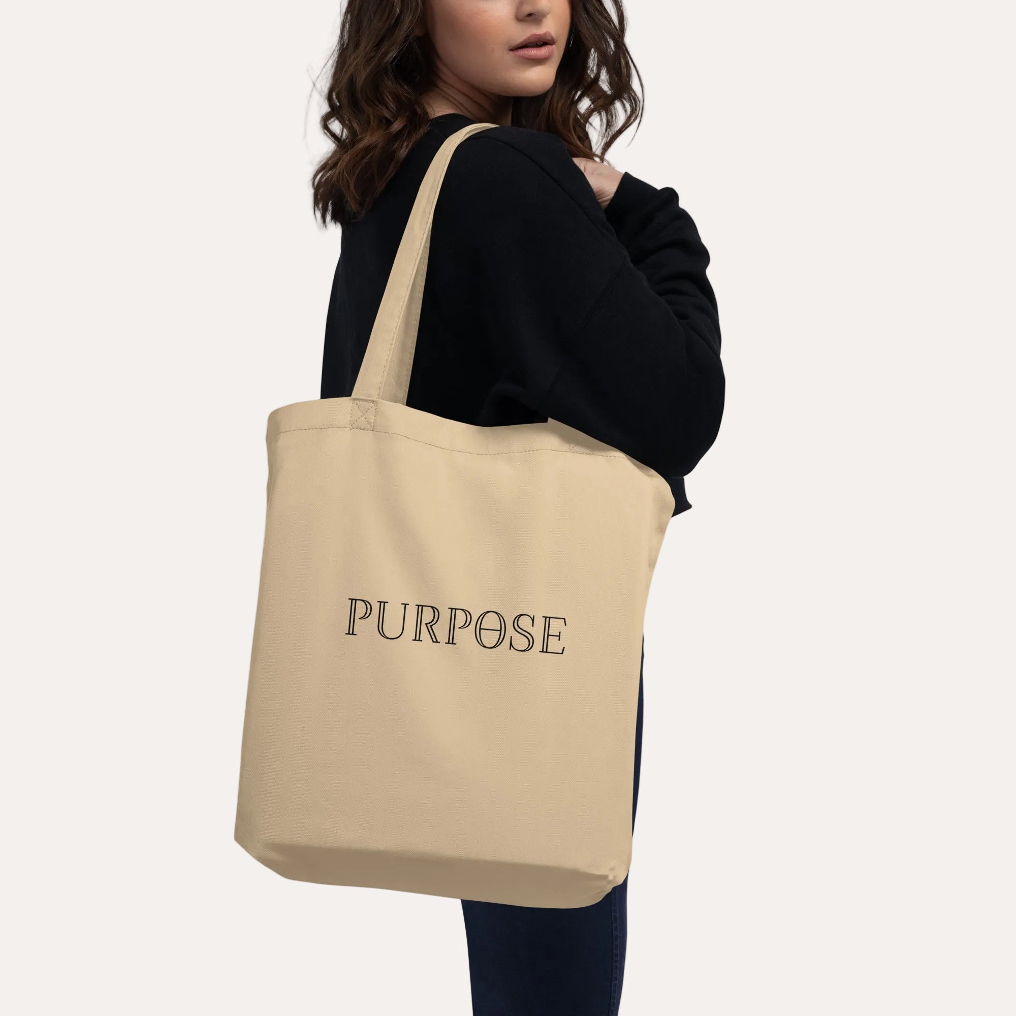 A woman wearing a black sweatshirt and jeans carries a black canvas tote bag over her shoulder. The bag features the word 'PURPOSE' printed in white serif font on the back, set against a plain white background.