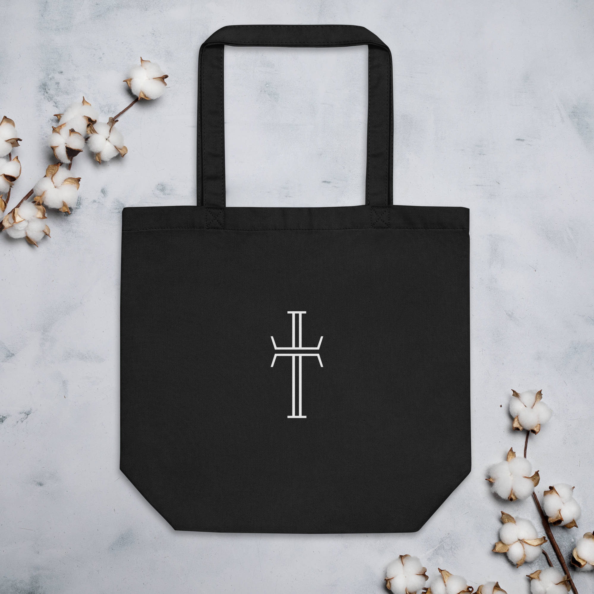 Black cotton canvas tote bag with shoulder straps and a centered white stylized cross on the front.
