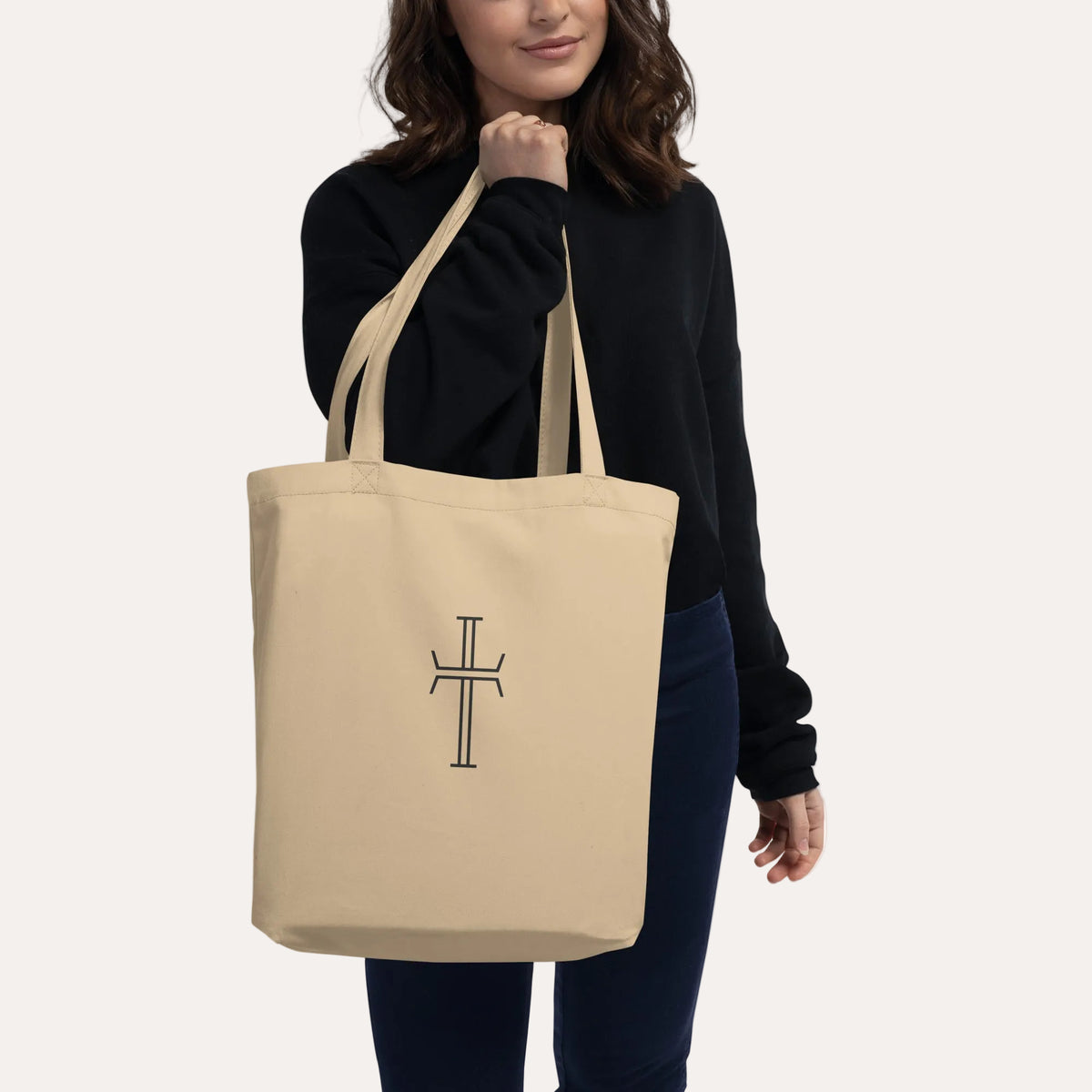 A woman wearing a black sweatshirt and jeans carries a natural canvas tote bag in her hand. The bag features the Jesus Cross on the front, set against a plain white background.