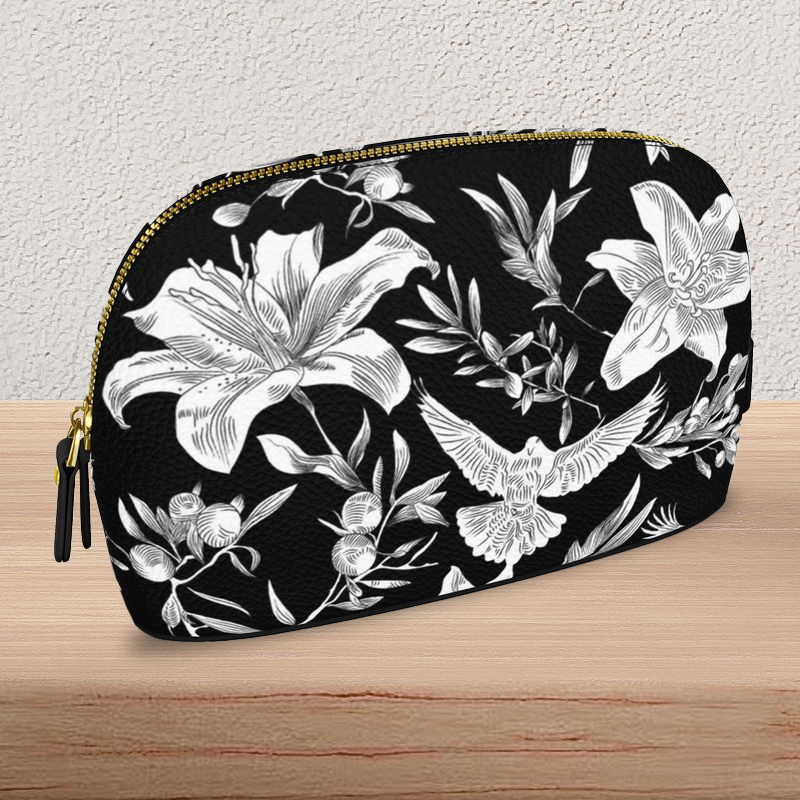 Angled close-up view of a large, black, Nappa-textured leather makeup bag with gold-colored zipper and pulls, sits on a light-colored wooden console table. The bag is flat on the bottom with a curved top edge and has a bold white pattern of lilies, olive branches, and doves in an all-over print.